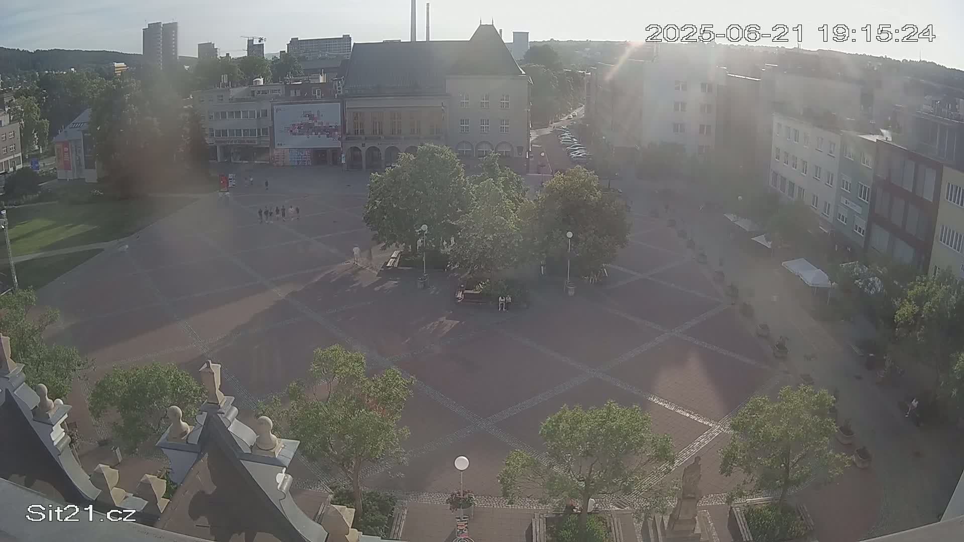 A high-angle, hazy view of a town square on a partly sunny day shows several buildings surrounding a paved area with small trees and a few people.