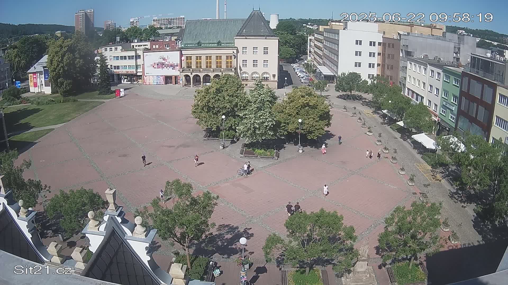 A large, paved town square with a few people walking and sitting, surrounded by buildings and trees under a sunny sky.