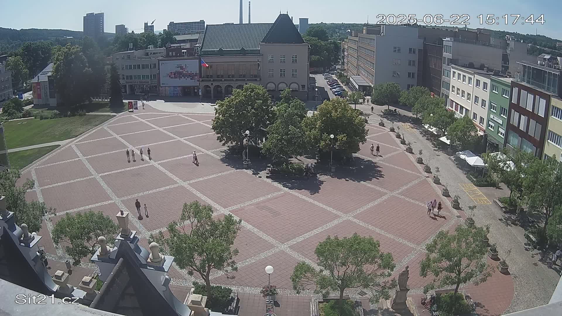 A large, brick town square with scattered pedestrians and trees under a sunny sky.