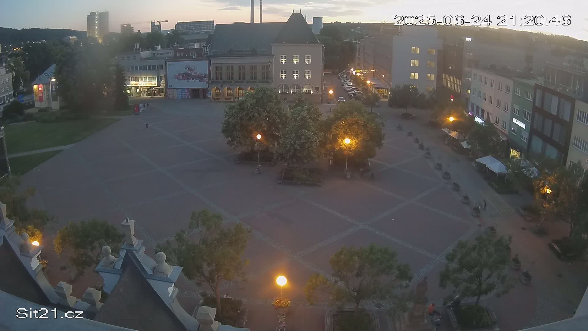 An empty town square at dusk, illuminated by streetlights, is surrounded by buildings and trees.