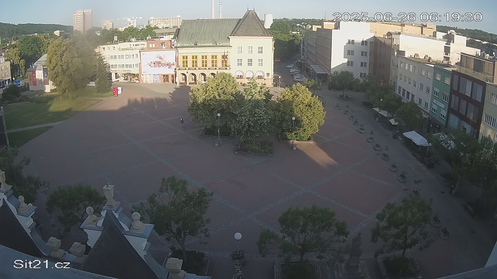 An empty town square at dusk, illuminated by streetlights, is surrounded by buildings and trees.