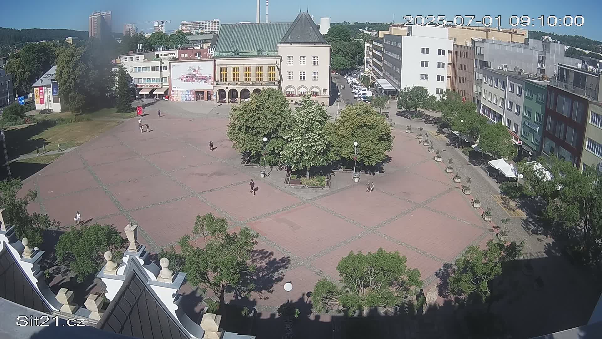 A paved town square with several buildings surrounding it, a few people walking around, and some trees planted in the center, on a sunny day.