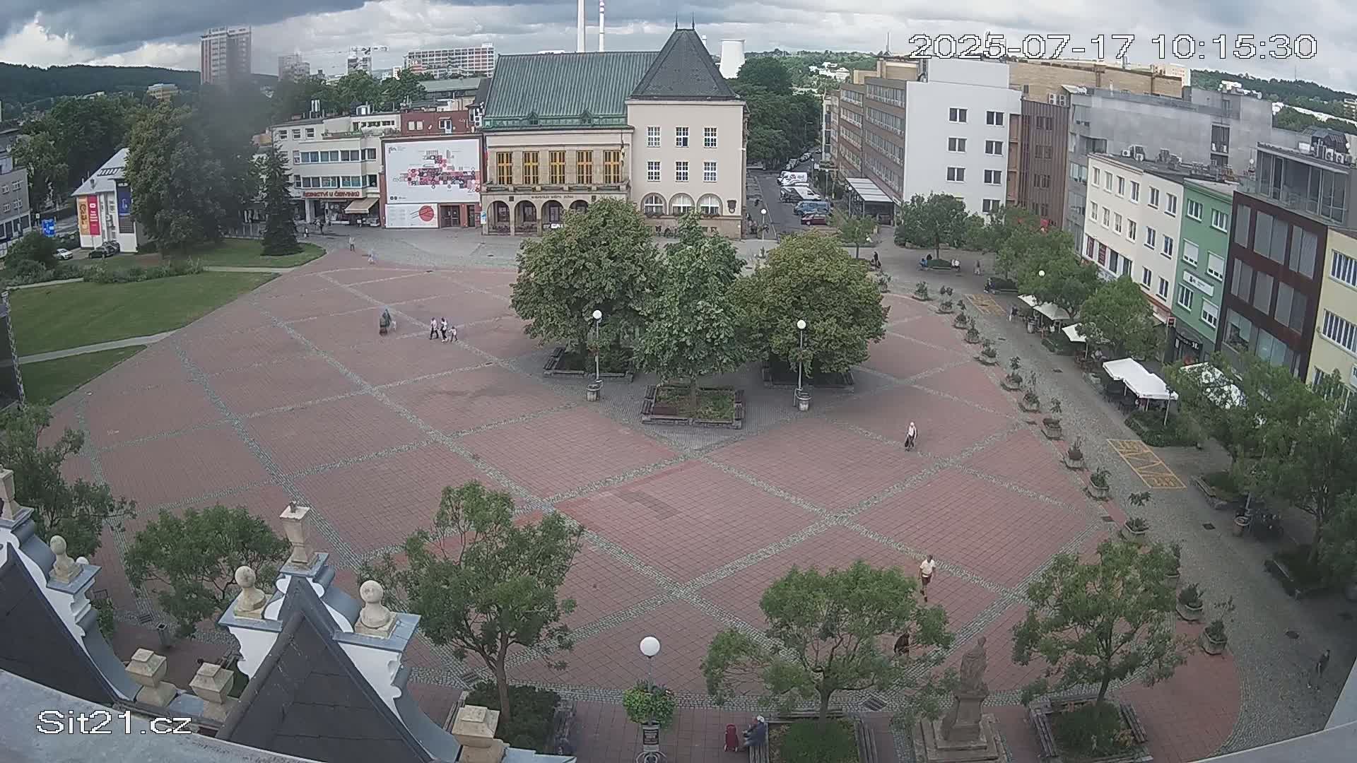 An overhead view of a town square with brick paving, several trees, and a few people walking around under cloudy skies.
