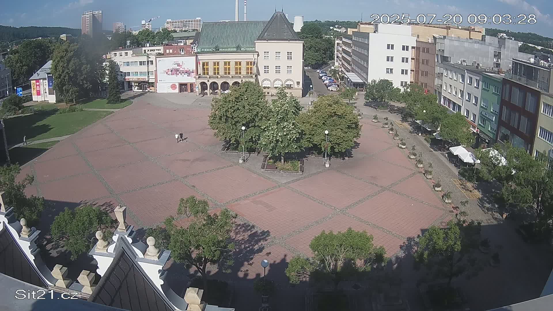A mostly empty, brick-paved town square is surrounded by buildings and trees under a sunny sky.