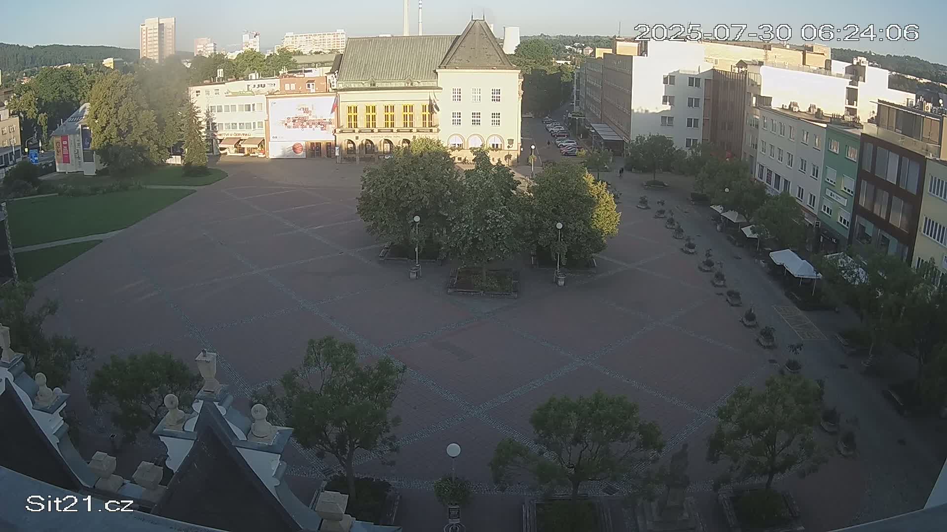 A mostly empty, brick-paved town square is surrounded by buildings and trees under a sunny sky.