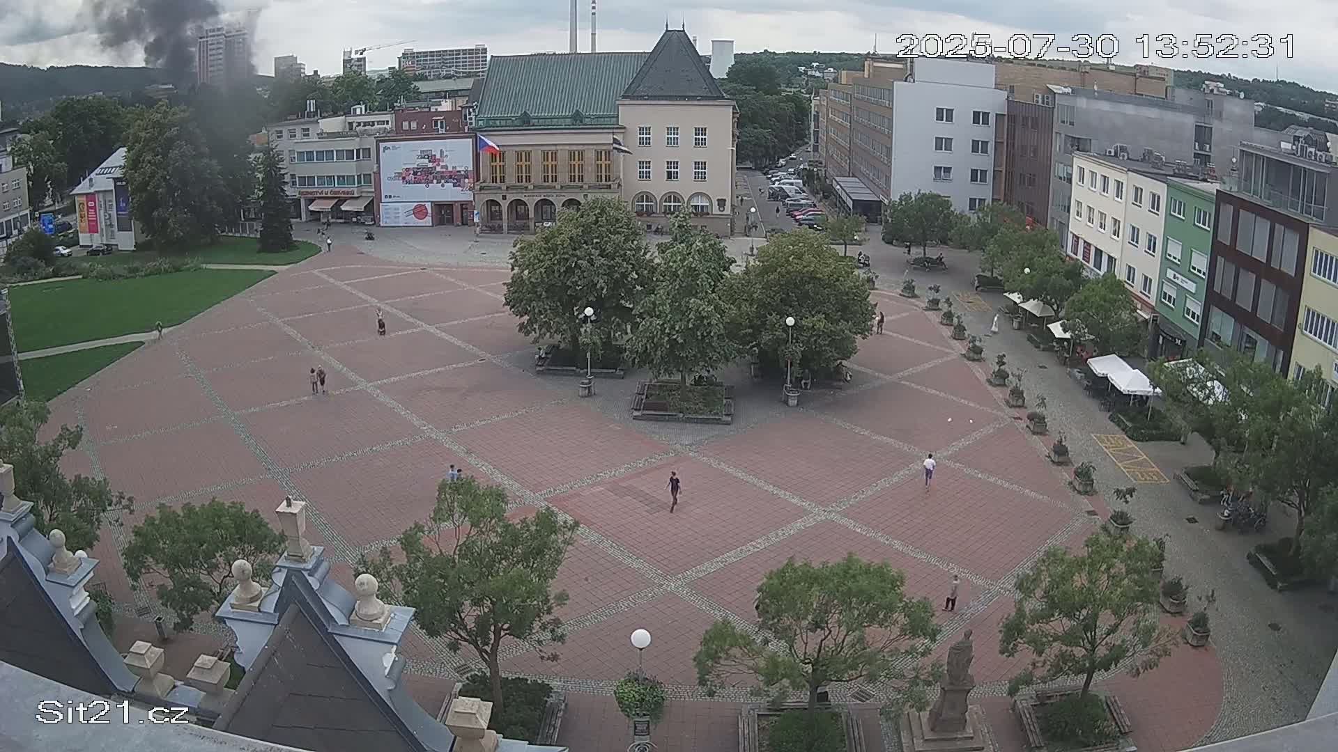 A mostly empty, brick-paved town square is surrounded by buildings and trees under a sunny sky.