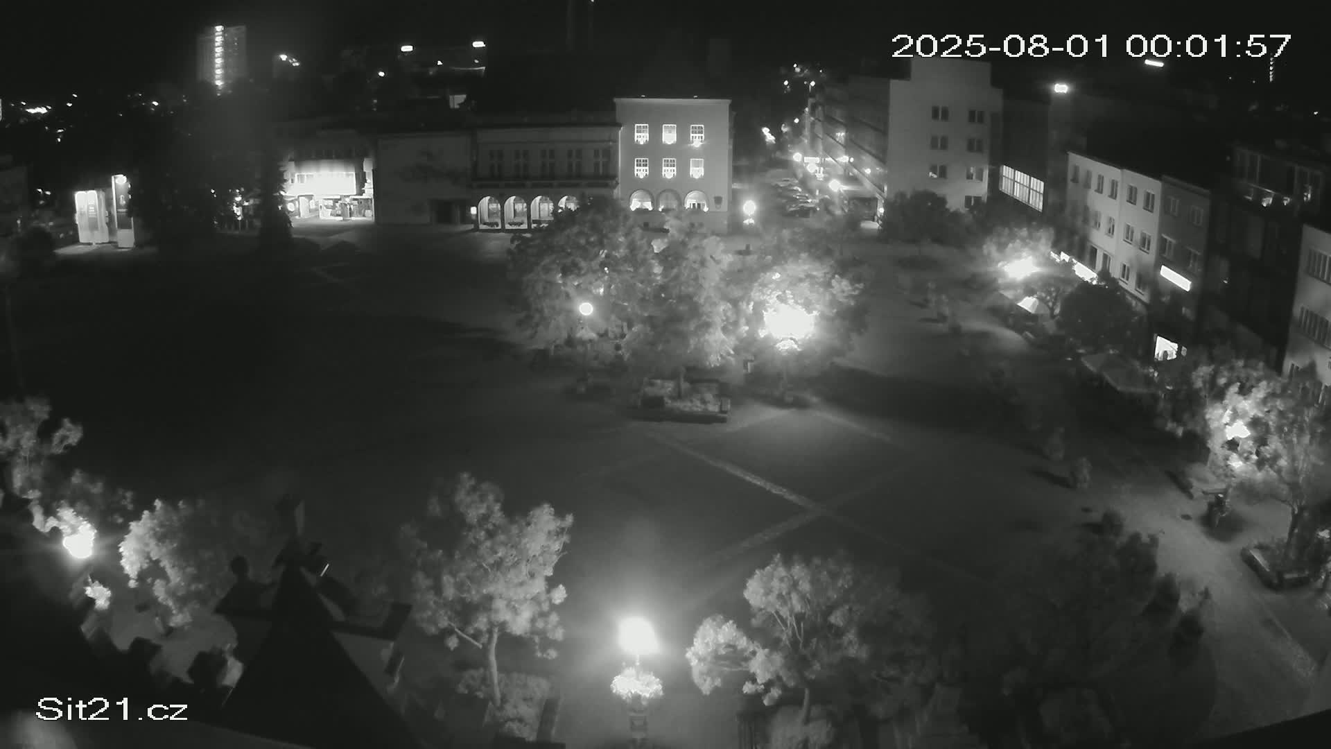 A nighttime, high-angle view of a town square with buildings surrounding a paved area containing trees and lampposts.