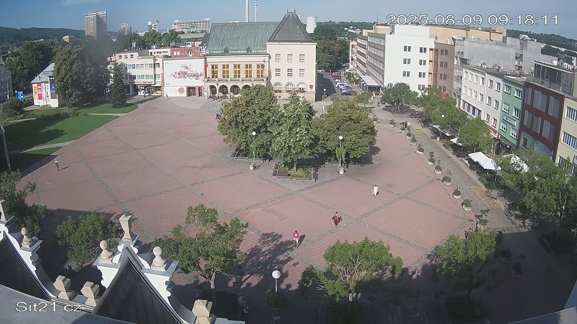 An elevated view shows a town square paved with reddish-brown tiles, featuring a central garden with trees, surrounded by buildings and people walking on a sunny day.