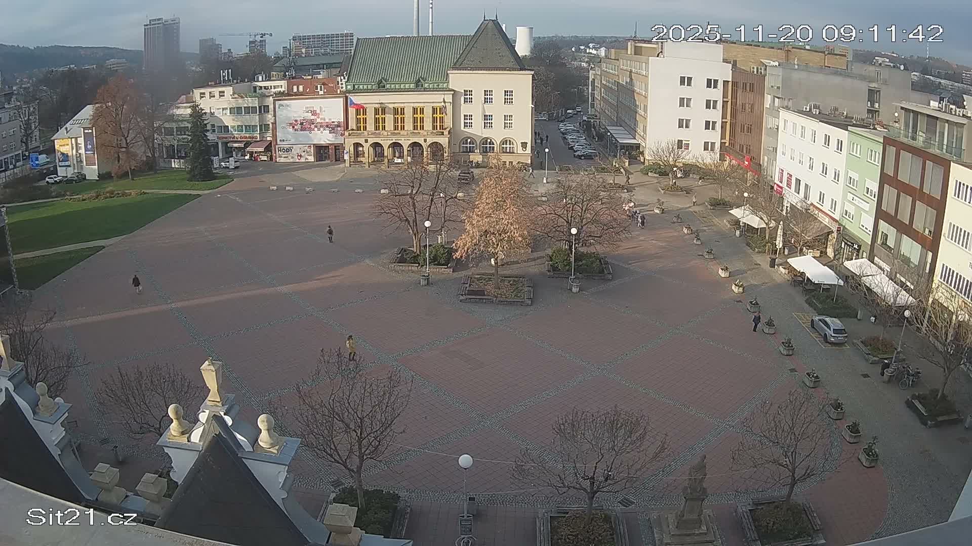 An elevated view shows a city square bustling with pedestrians, featuring mostly bare trees and surrounding buildings, all beneath a bright, partly cloudy sky.