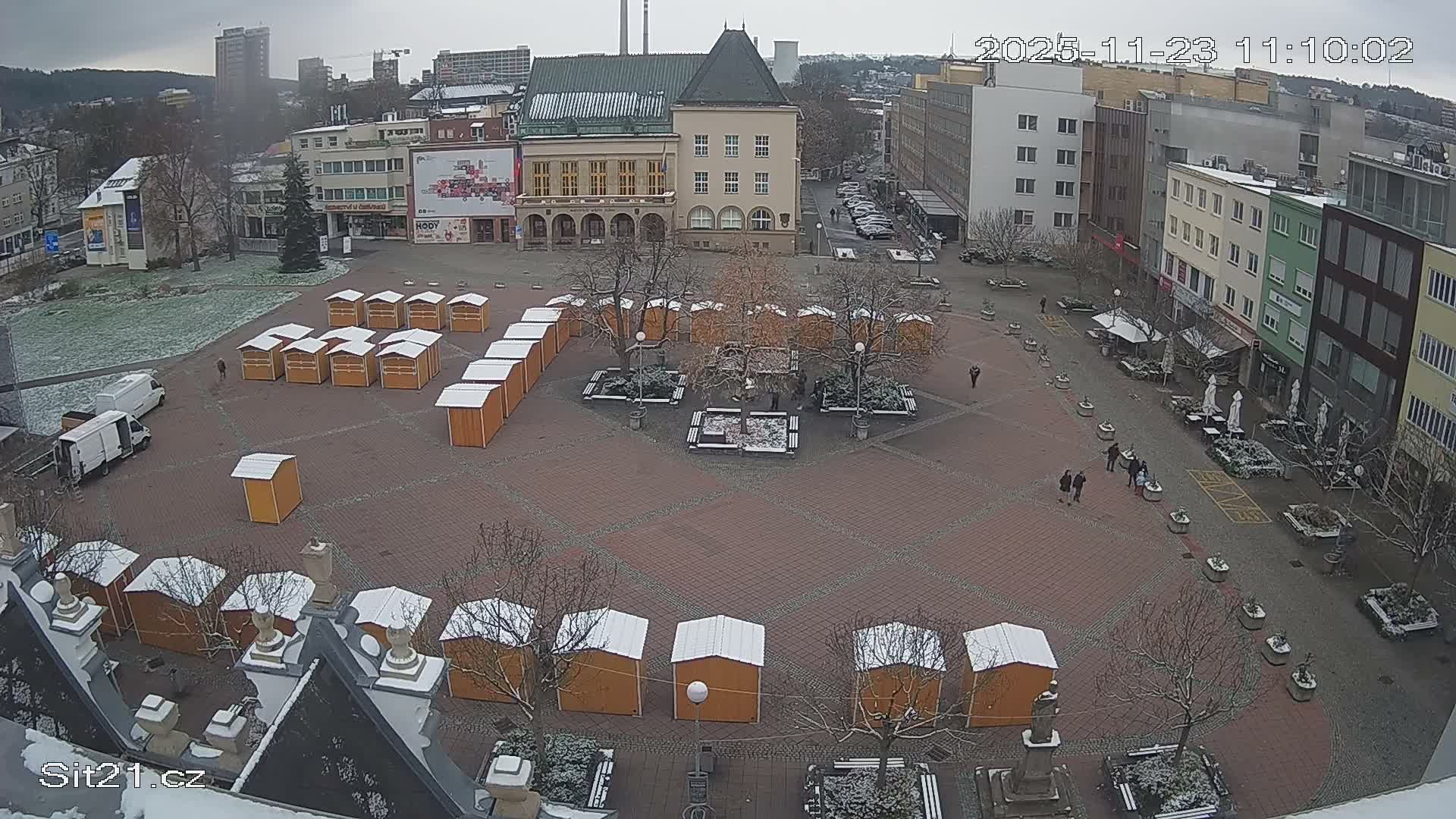 A sprawling urban square is lightly dusted with snow on an overcast winter day, filled with numerous wooden market stalls, bare trees, and people walking amidst surrounding buildings.