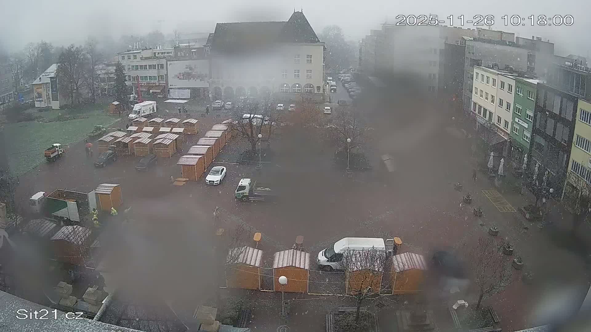 An elevated view captures a town square undergoing preparations, with numerous wooden market stalls being set up amidst parked vehicles and a few people, all under falling snow and an overcast sky.