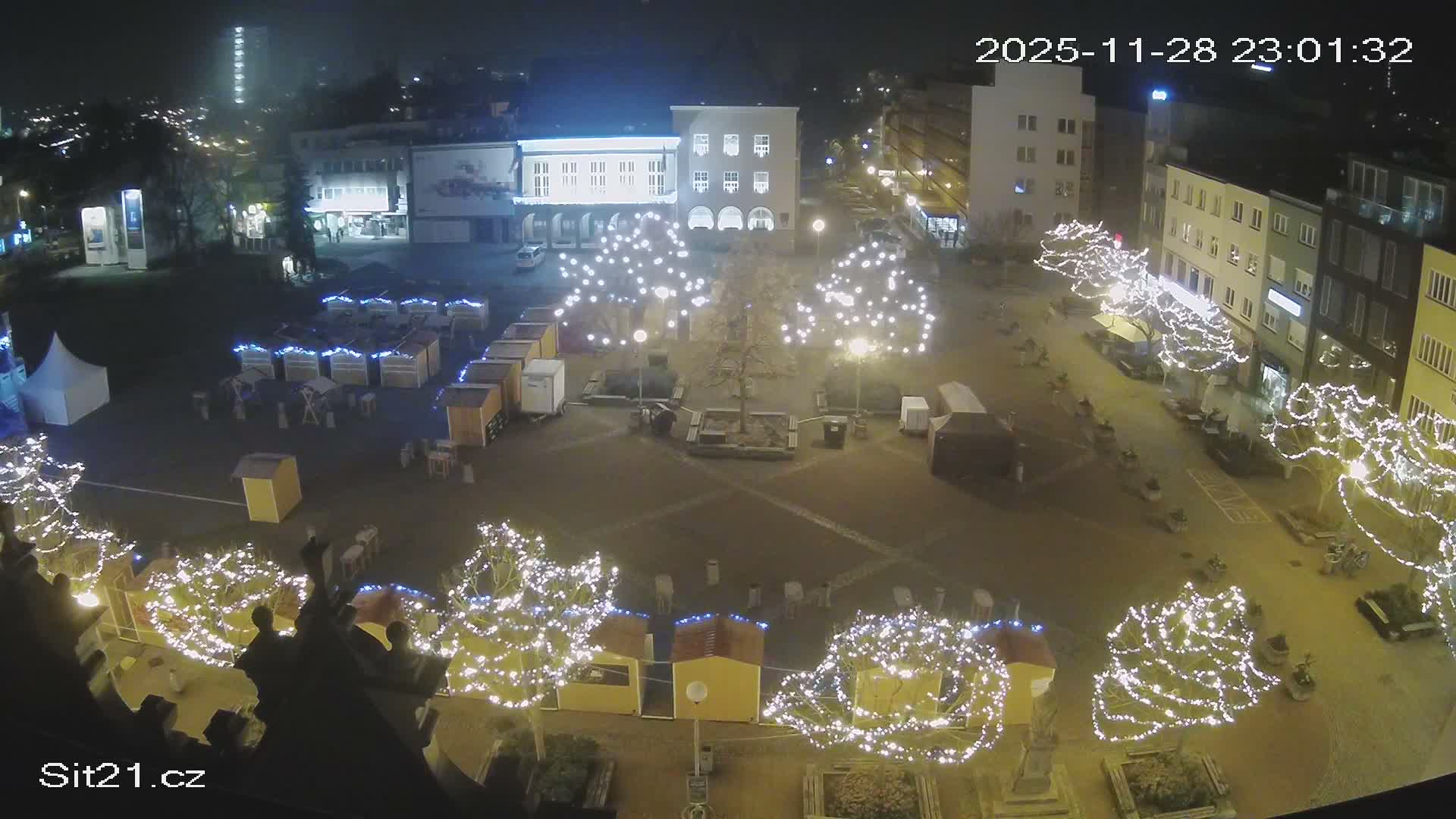 A festive outdoor market square is brightly illuminated by white string lights on trees and numerous stalls at night, under a clear sky.