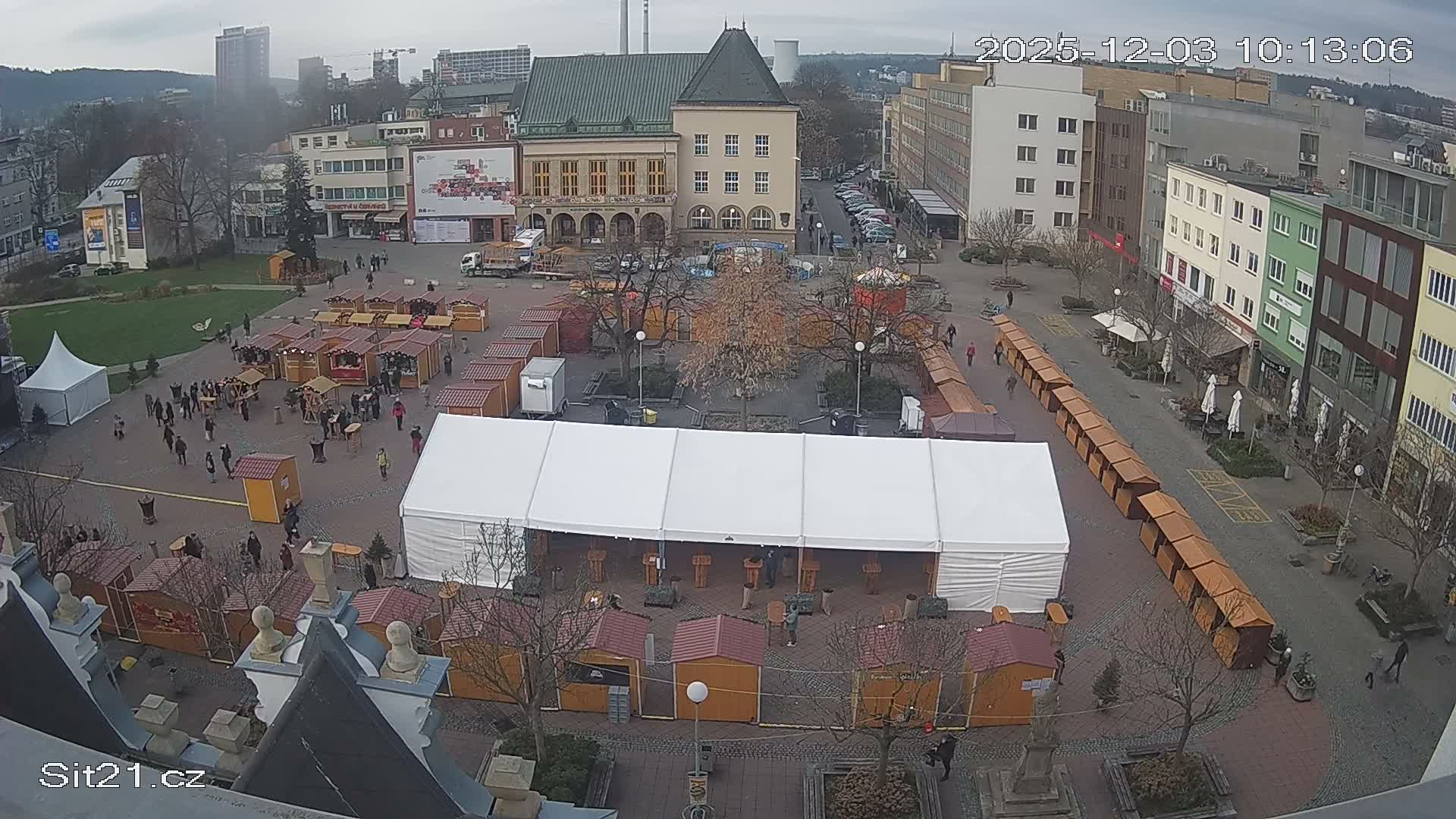 An elevated view reveals a bustling city square densely populated with numerous wooden market stalls and a large white event tent, as people mill about under a grey, overcast sky, surrounded by various city buildings including a prominent green-roofed structure.