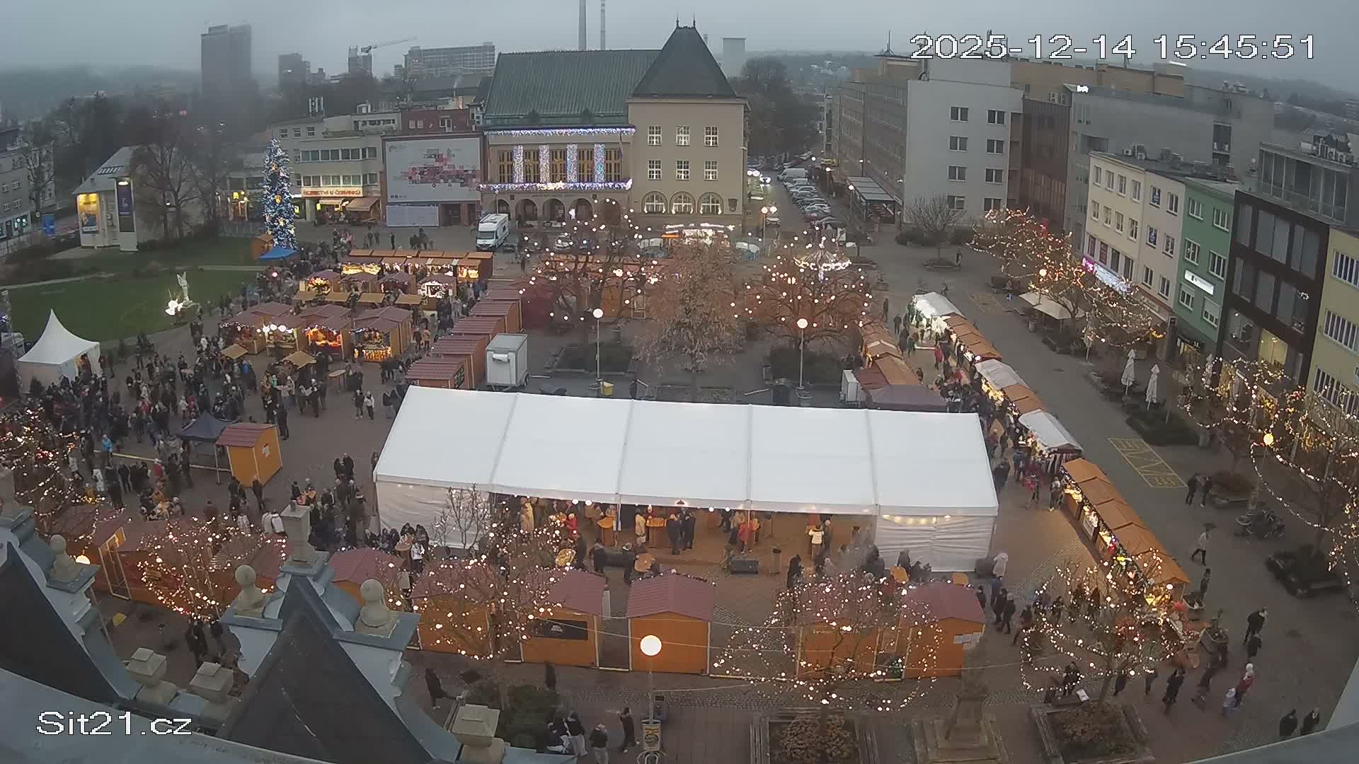 An elevated view reveals a bustling city square densely populated with numerous wooden market stalls and a large white event tent, as people mill about under a grey, overcast sky, surrounded by various city buildings including a prominent green-roofed structure.