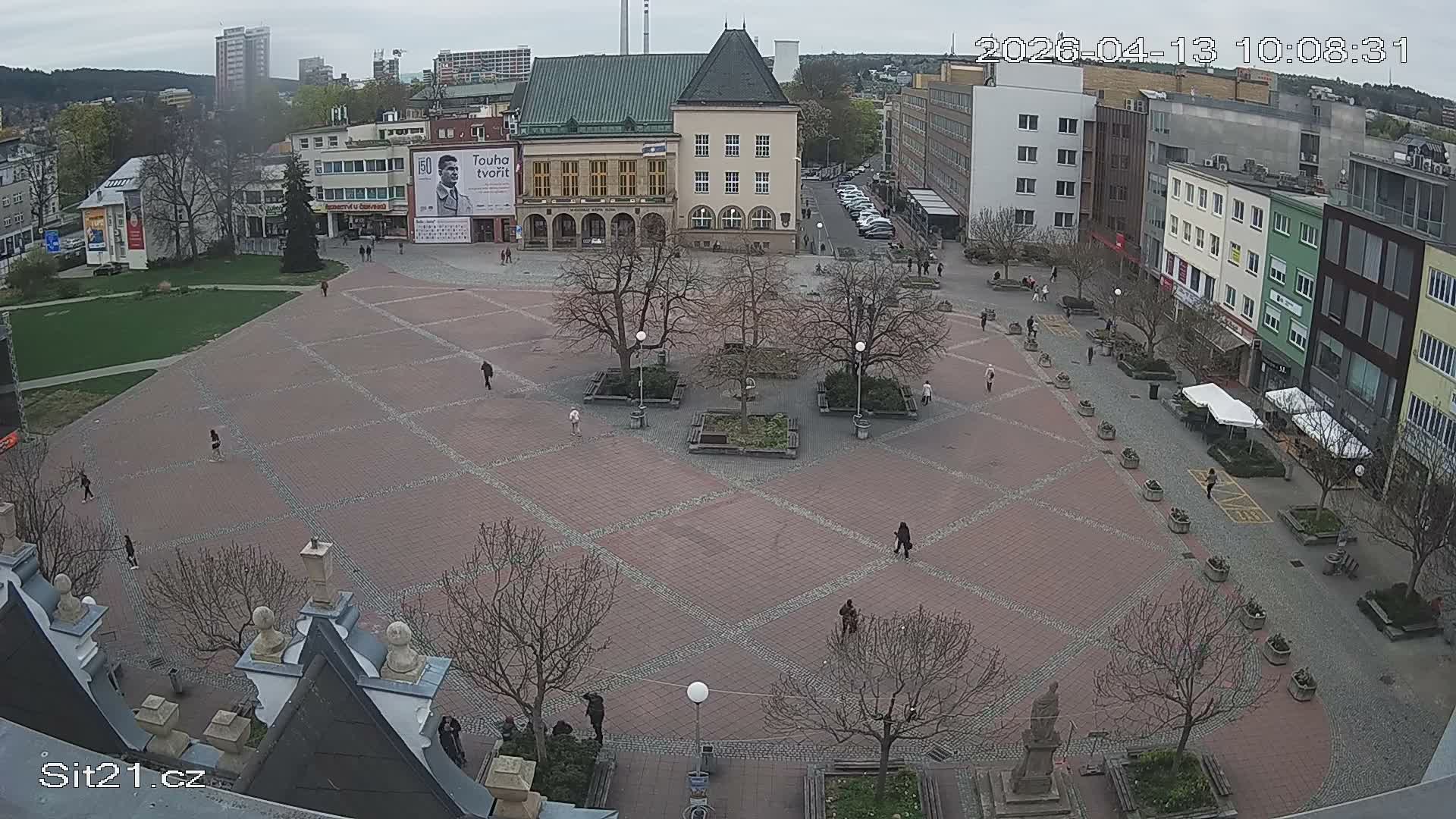 An elevated view reveals a bustling city square densely populated with numerous wooden market stalls and a large white event tent, as people mill about under a grey, overcast sky, surrounded by various city buildings including a prominent green-roofed structure.