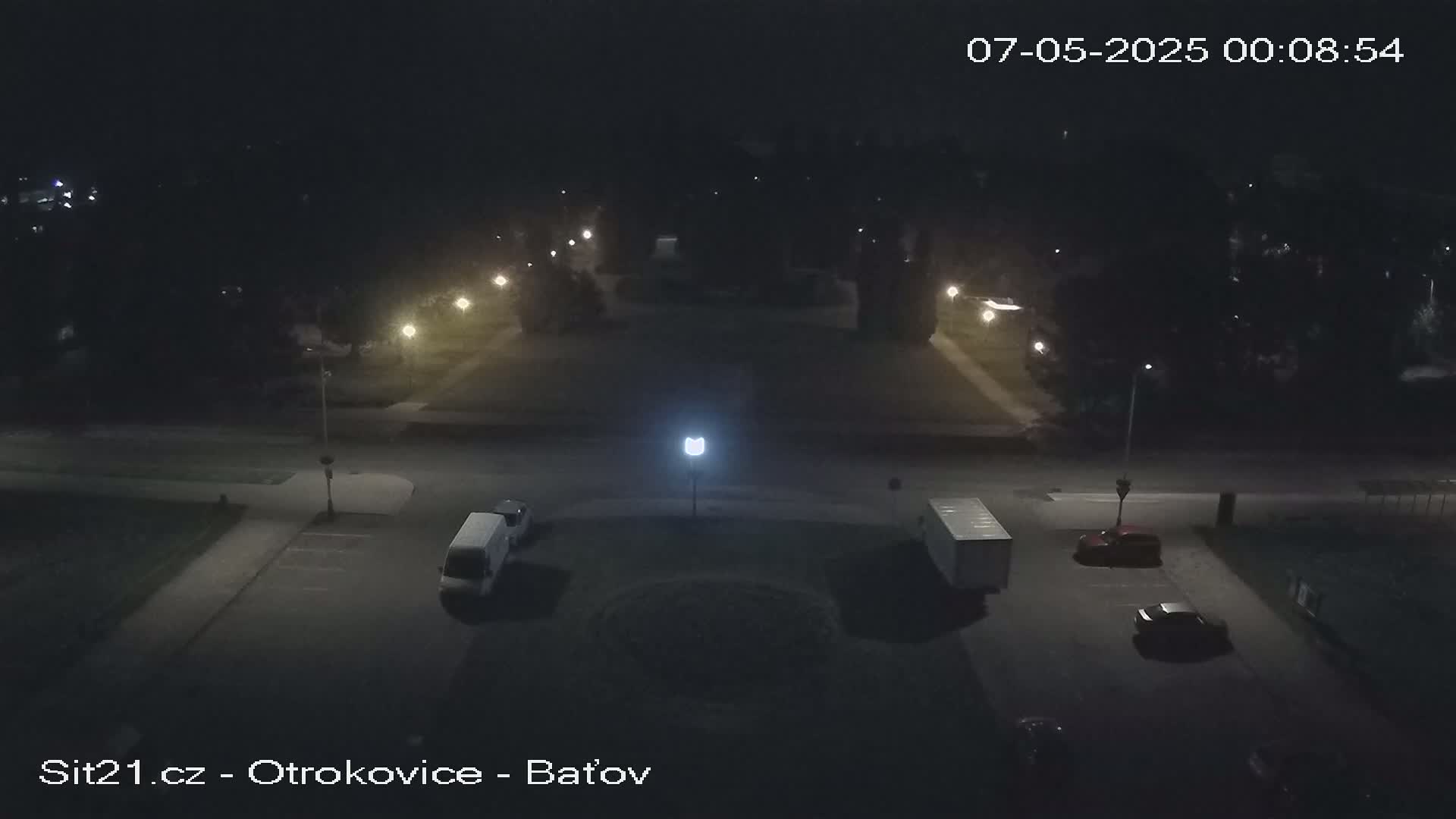 A nighttime aerial view of a mostly empty parking lot and street, sparsely lit by street lamps, with a few vehicles present.