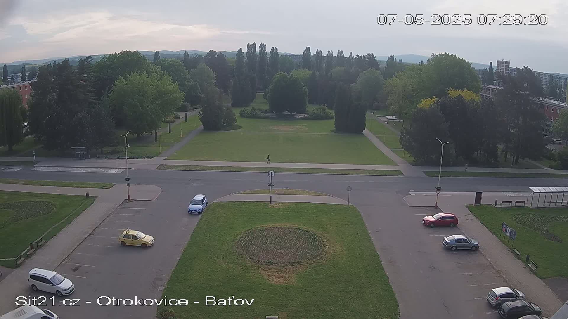 A high-angle view shows a park with a grassy area, surrounded by trees and buildings under an overcast sky, with several cars parked in a nearby lot.
