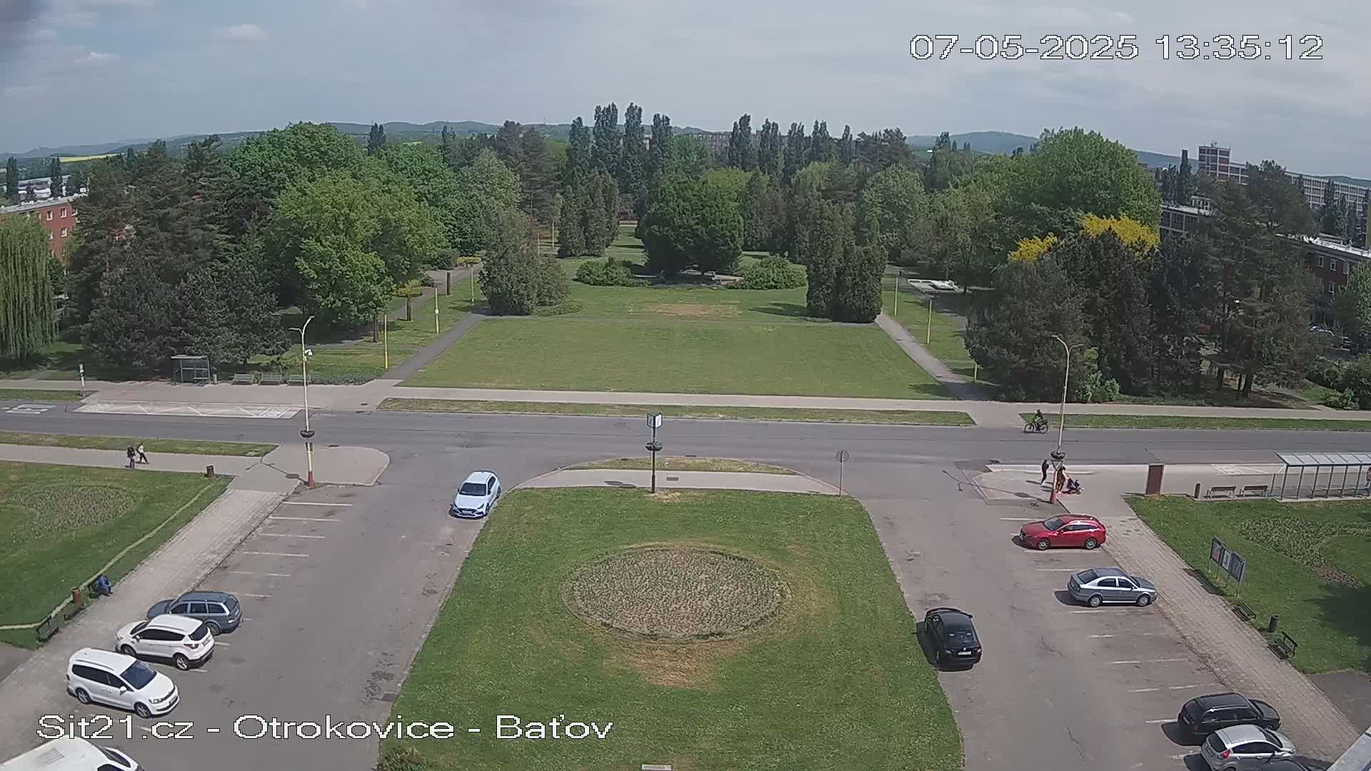 A high-angle view shows a park with several cars parked in a lot, surrounded by buildings and trees under a partly sunny sky.