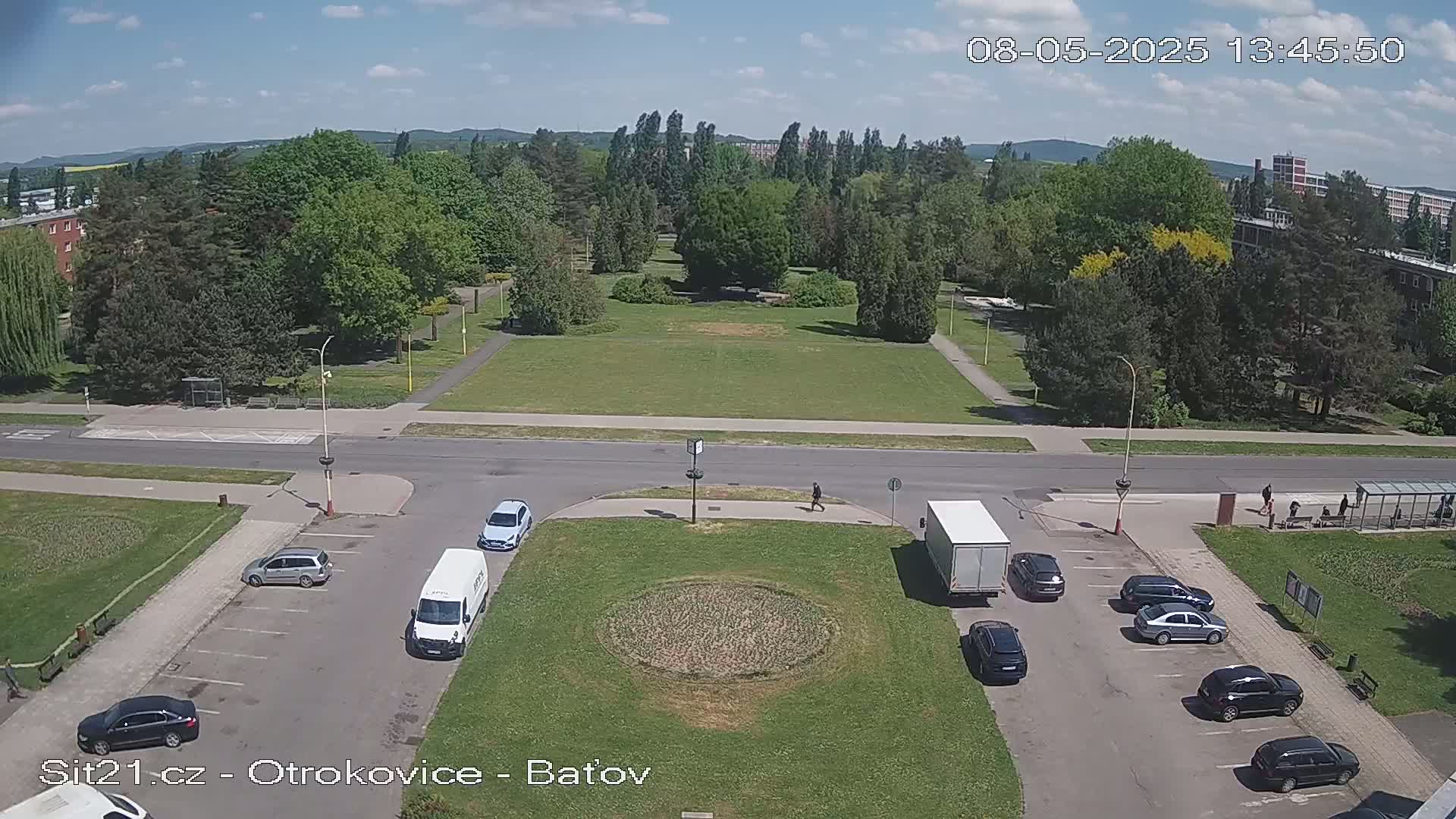 A high-angle view shows a sunny day with a green park, a roadway, parking lots with several cars and a van, and a white truck.