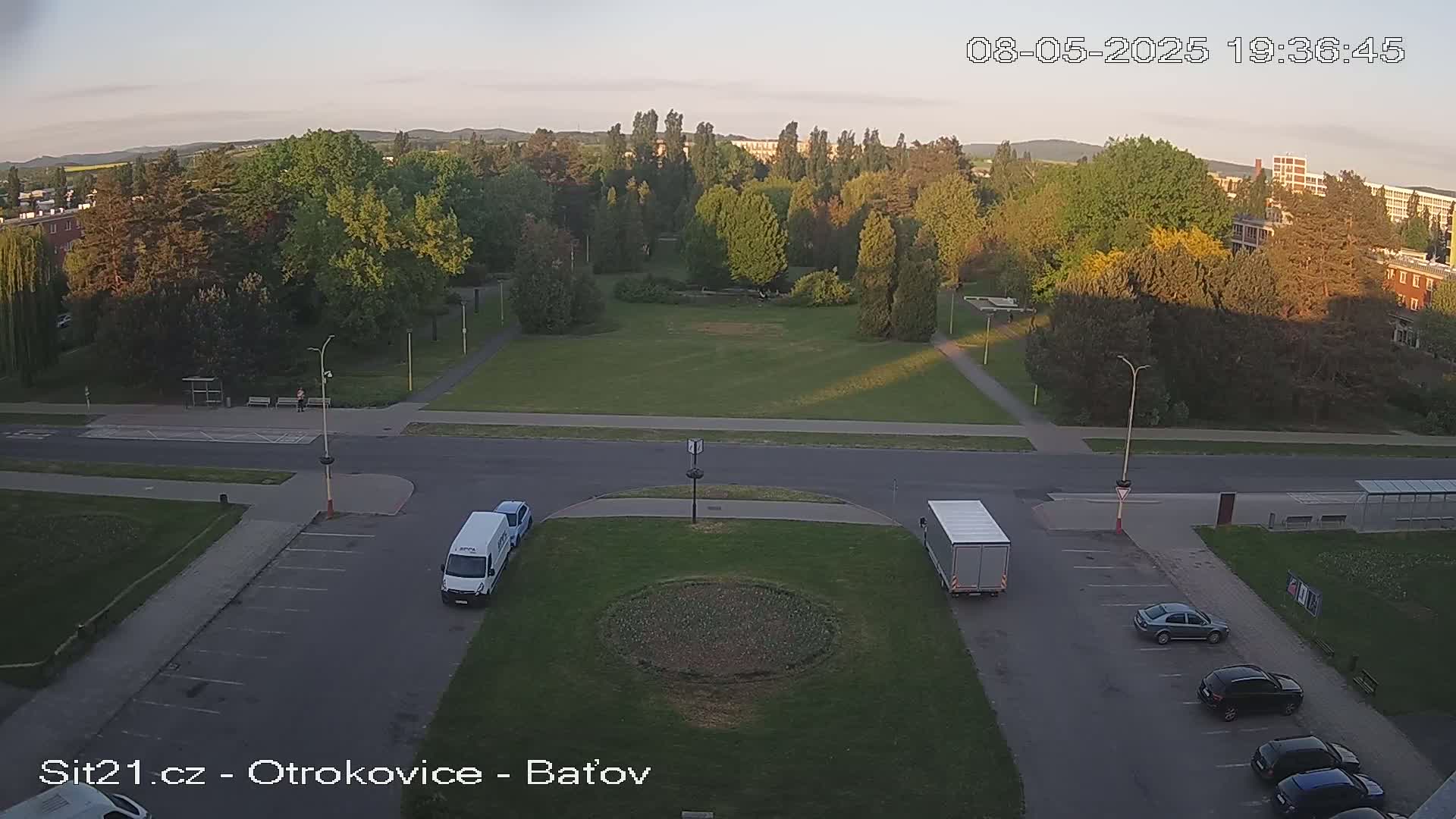 An elevated view of a park and parking lot with several cars and two vans, under a clear evening sky.