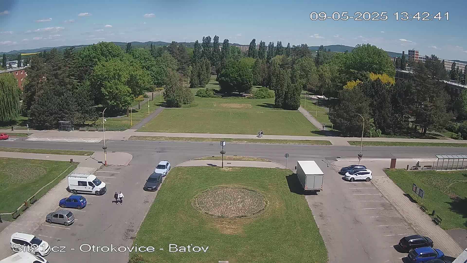 A sunny day view of a park and parking lot with several cars, a van, and a truck, situated near a road.