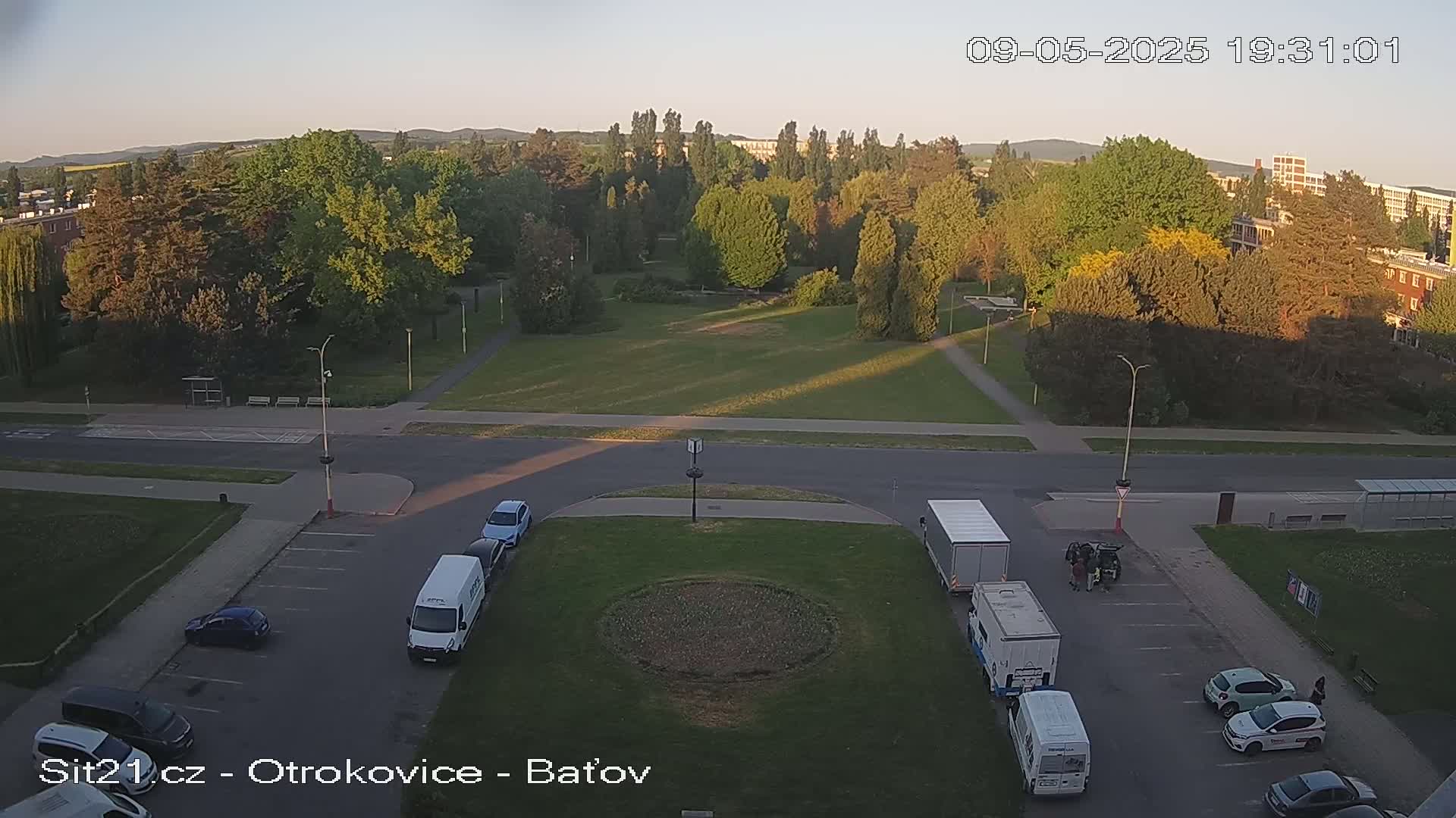 An elevated view of a park and surrounding area at sunset, showing several vehicles parked in a lot, with some vans and trucks in the parking area.