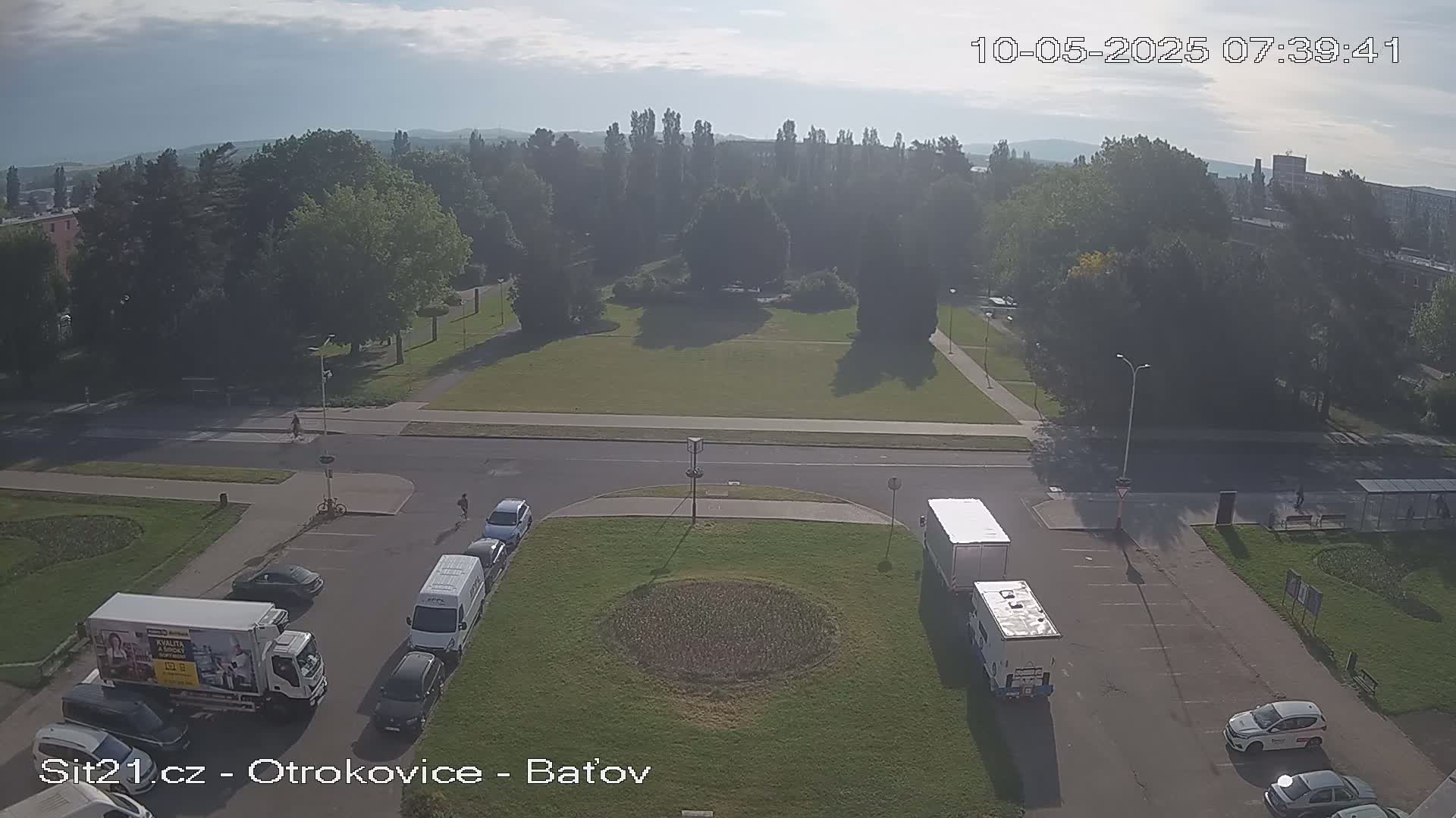 A hazy, sunny day shows a high-angle view of a park and roads with several vehicles parked and some pedestrians, including a bicycle, near a large grassy area with a circular patch in the center.