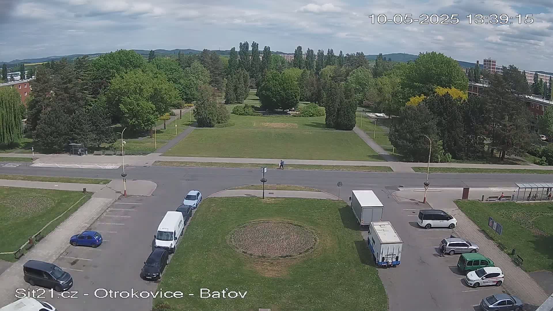A parking lot with several cars and trucks is situated next to a park with lush green grass and trees under a partly cloudy sky.