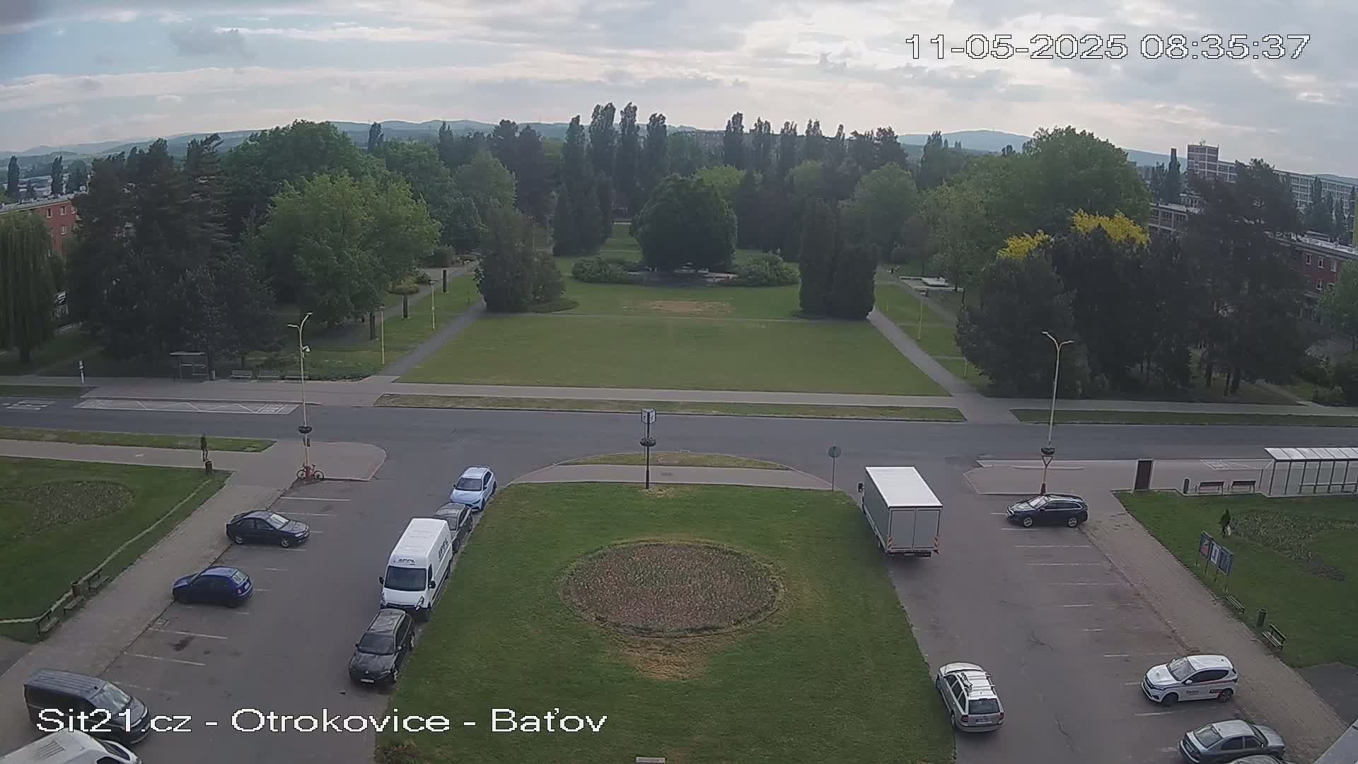 An overcast day reveals a high-angle view of a park and surrounding streets, with several cars parked and a truck stopped at an intersection.