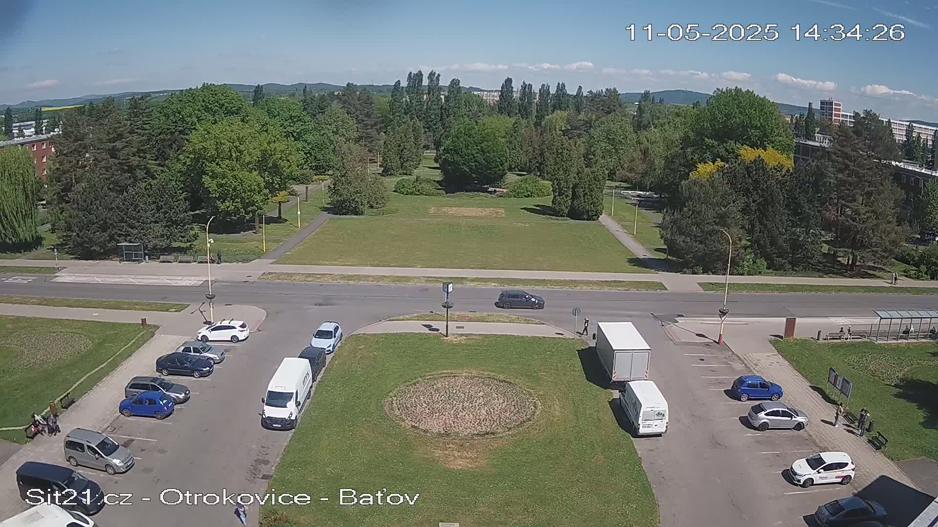 An aerial view shows a park with green grass and many trees, surrounded by a road with parked cars and vans on a sunny day.