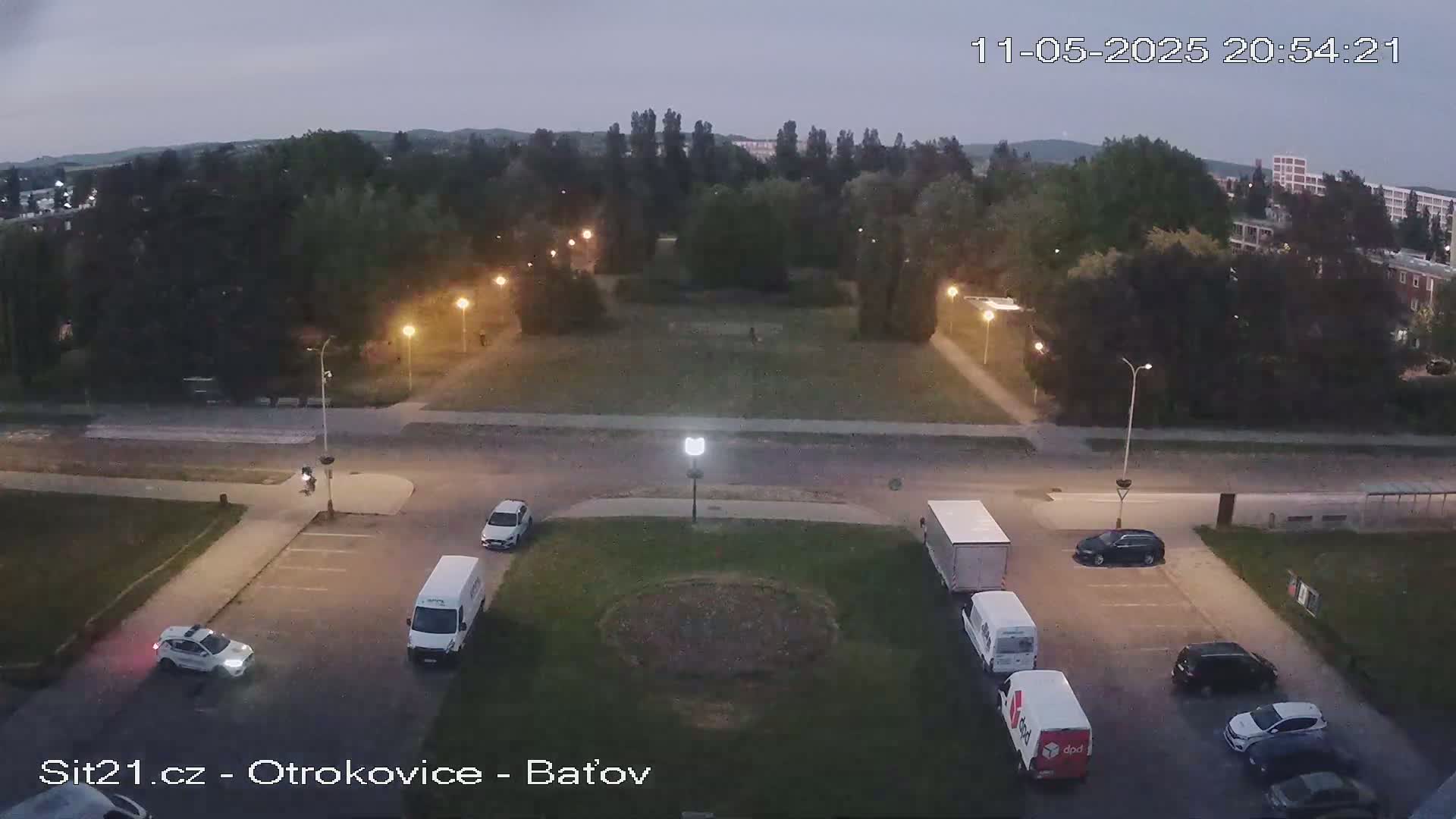 A twilight view of a park and surrounding streets with several vehicles parked and driving, under a cloudy sky.