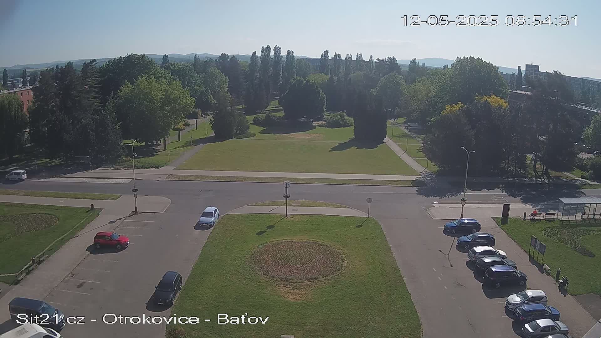 A high-angle view shows a park with a circular patch of low plants surrounded by a road with several cars parked along the sides, under a clear, sunny sky.