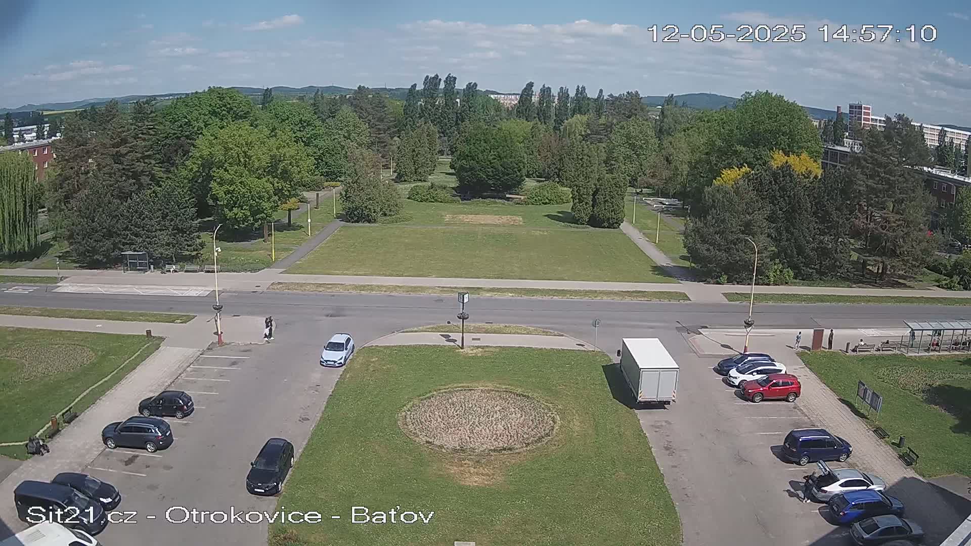 An overhead view shows a park with green grass and trees under a mostly sunny sky, with cars parked in a lot beside a roadway.