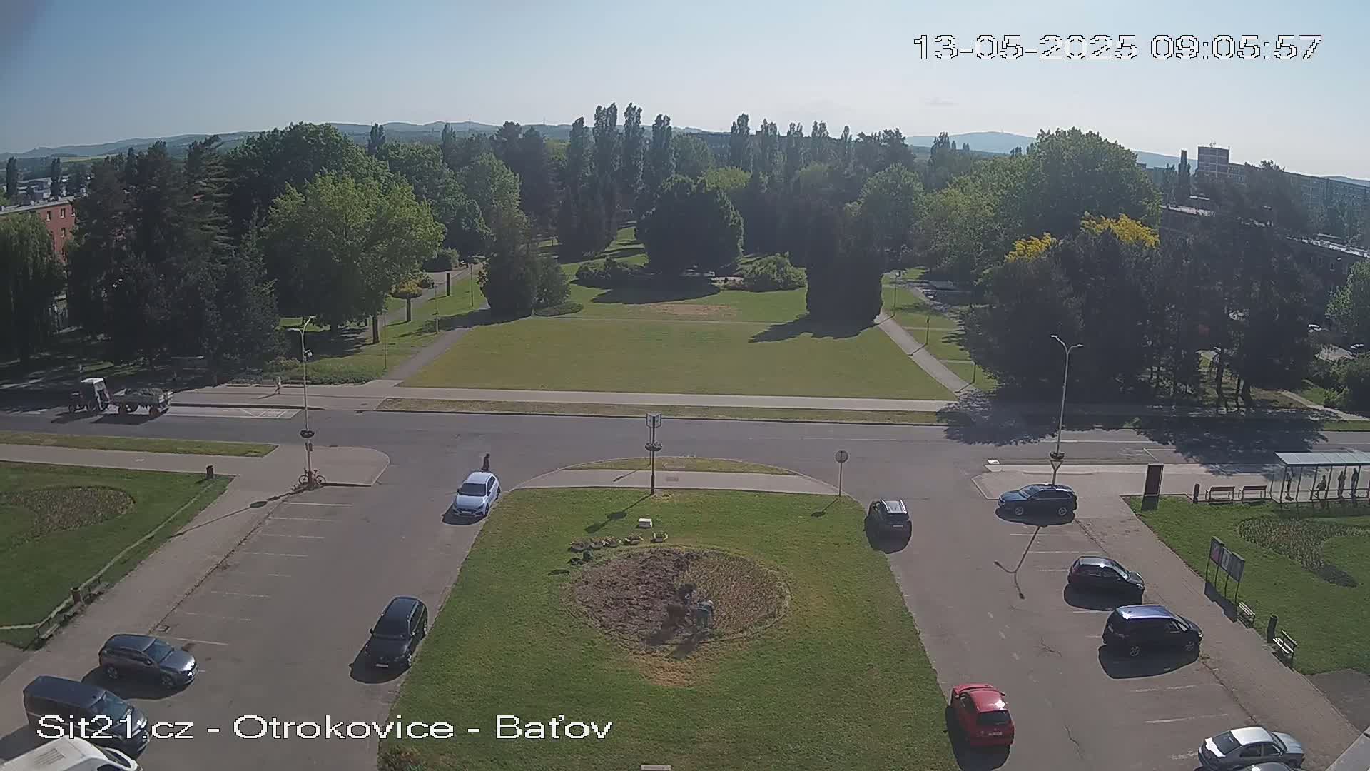A high-angle, sunny view of a park with a roadway and parking lot containing several cars, surrounded by trees and buildings.