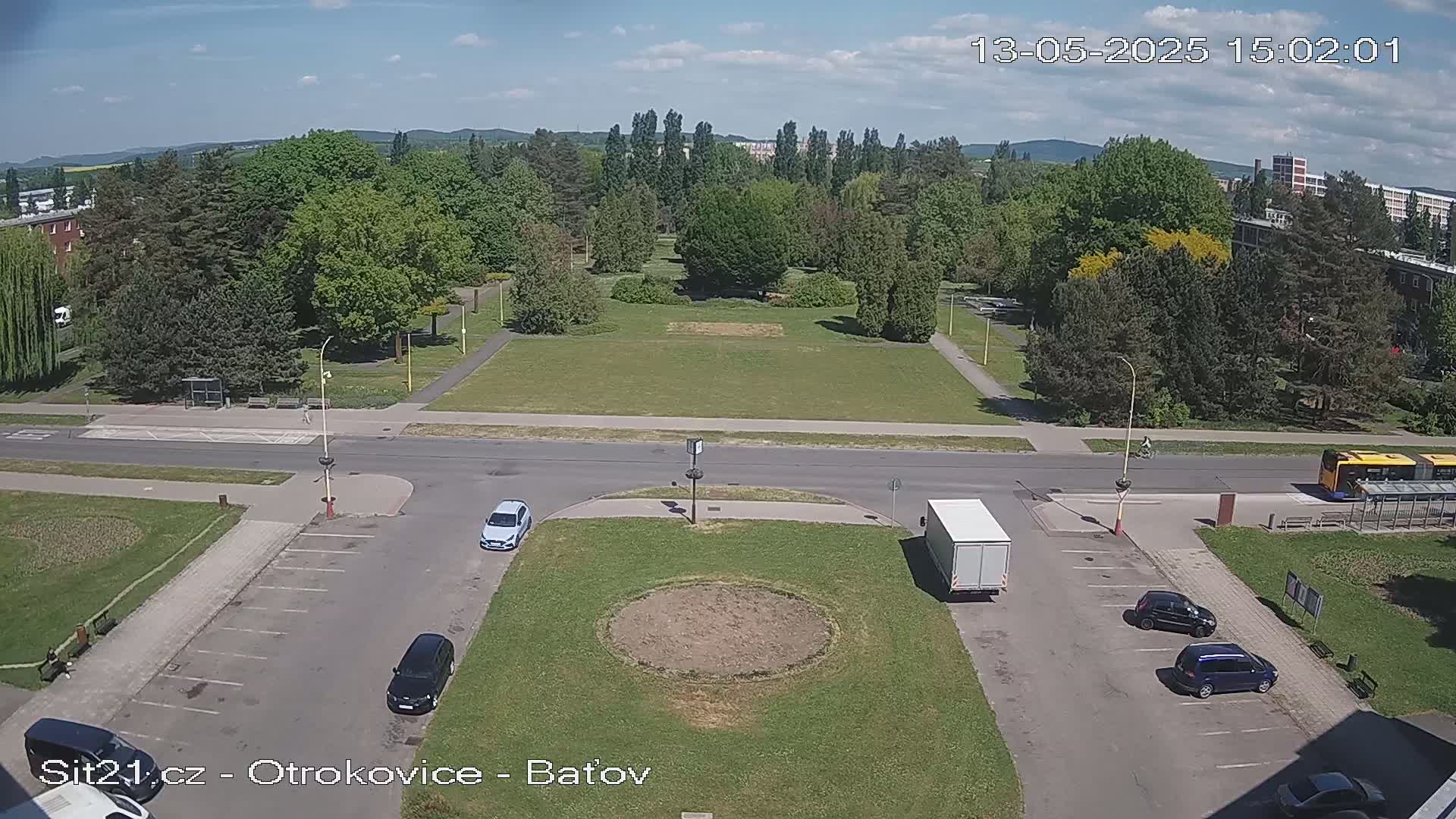 A sunny day reveals a high-angle view of a parking lot, a road, and a large grassy park with several trees, featuring several cars and a bus in the vicinity.