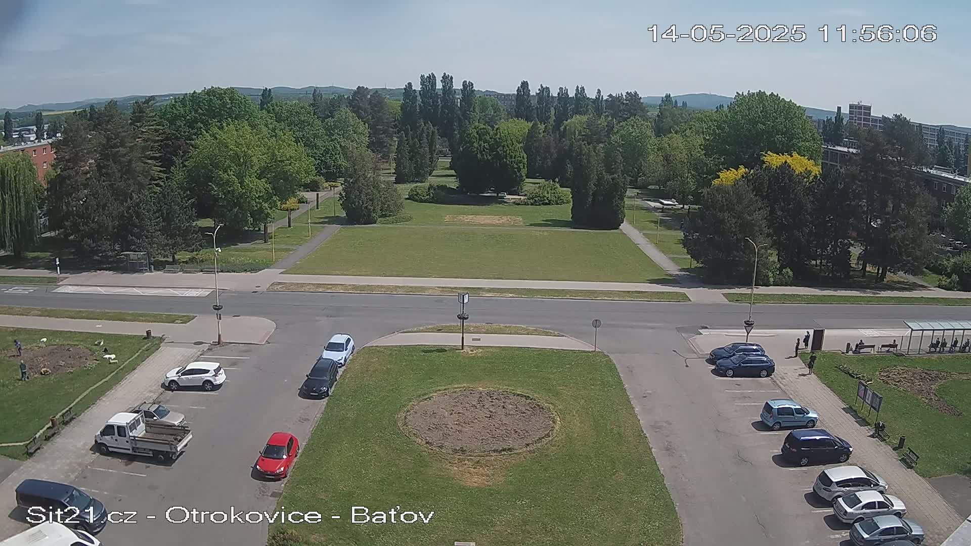 A sunny day reveals a high-angle view of a town square with a grassy area, surrounded by trees, buildings, and parked cars.
