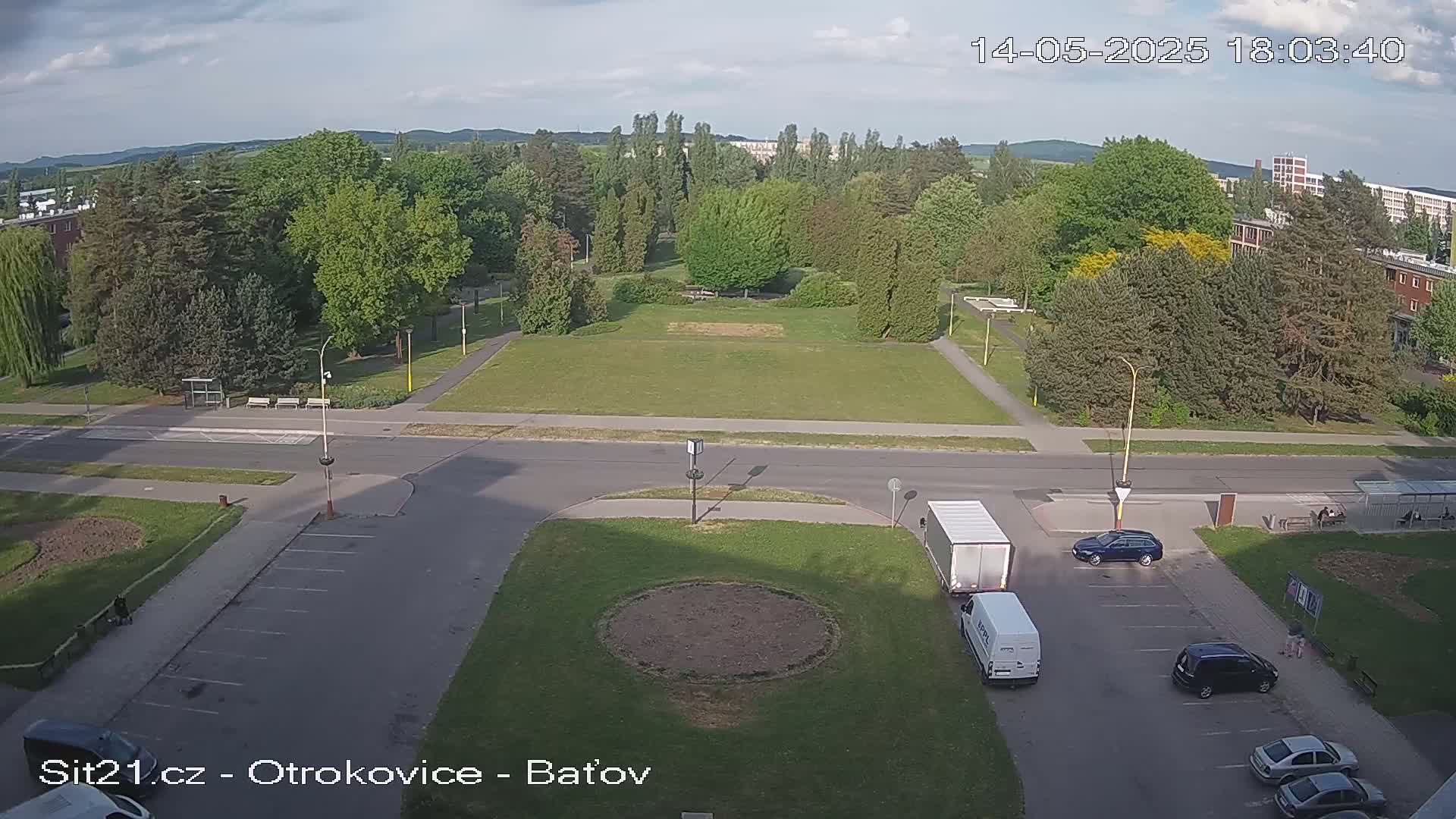 A mostly sunny high-angle view of a grassy park, a road intersection, and a parking lot with several vehicles, situated amongst buildings and trees.