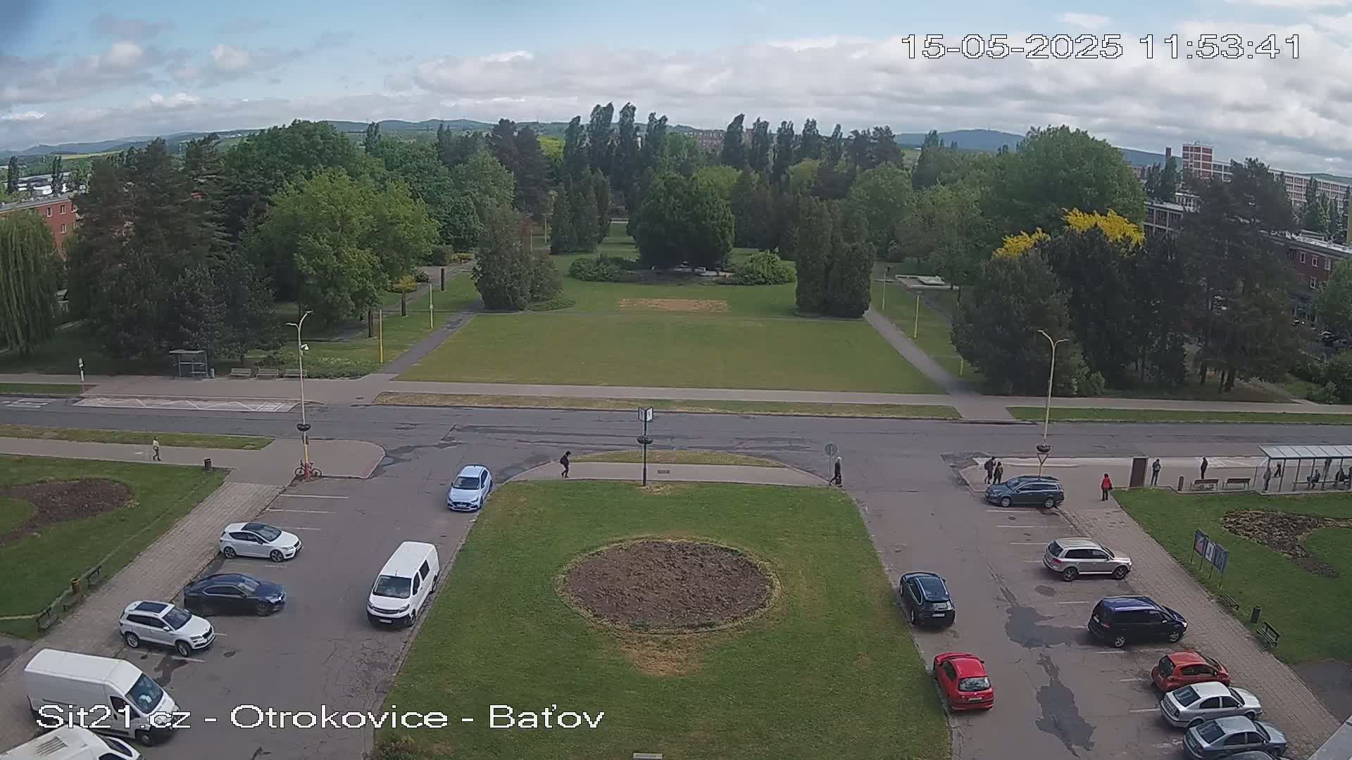 An elevated view shows a park with a large grassy area, surrounded by trees, buildings, and cars parked in a lot under a partly cloudy sky.