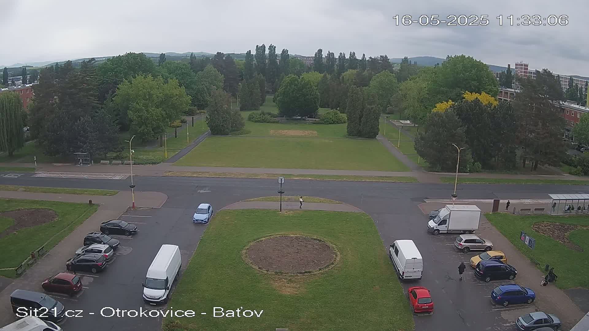 A high-angle view shows a parking lot with several cars and vans, a person walking in a grassy area, and a park with lush green trees under an overcast sky.