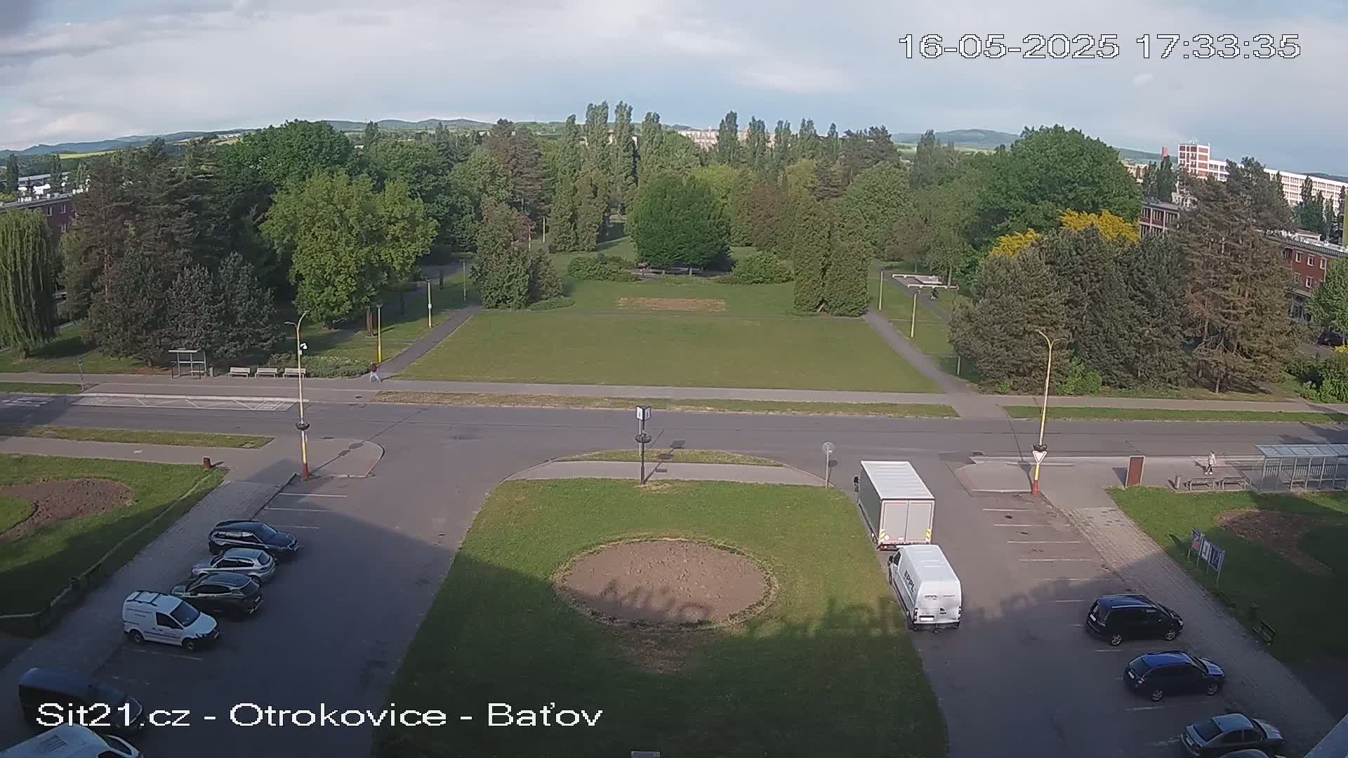 A mostly sunny overhead view shows a large grassy park bordered by trees and buildings, with a road running through it and several cars parked in a lot.