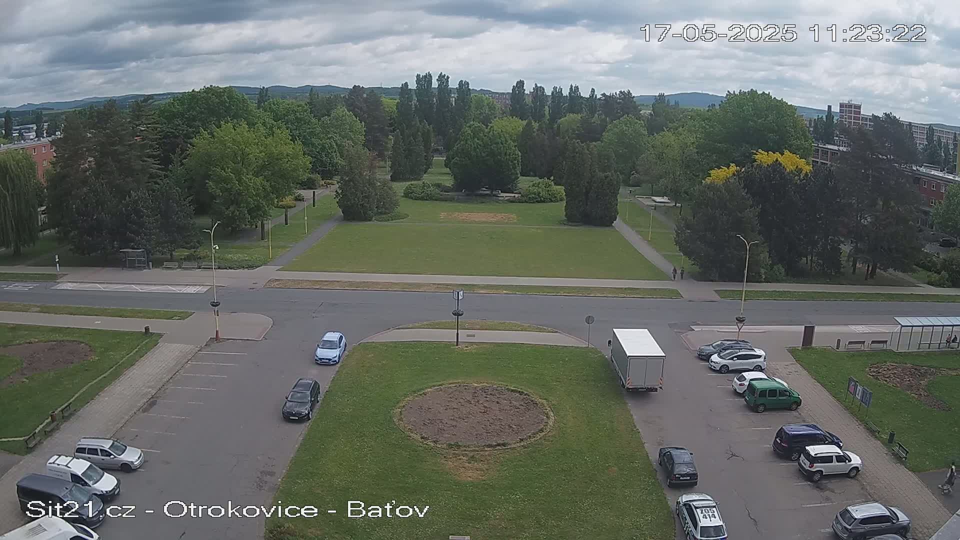An aerial view of a park with a roadway and parking lot, under overcast skies, shows several cars parked and a truck stopped in the lot.