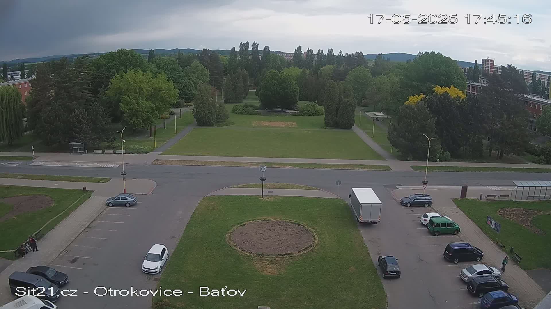 An overcast day shows a large grassy park surrounded by buildings and trees, with several cars parked in a lot next to a road.
