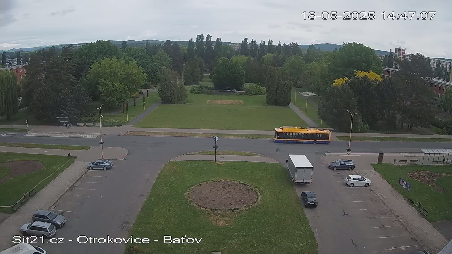 A high-angle view shows a street scene under overcast skies, with several cars parked near a grassy area, a yellow bus, and a white truck at an intersection.