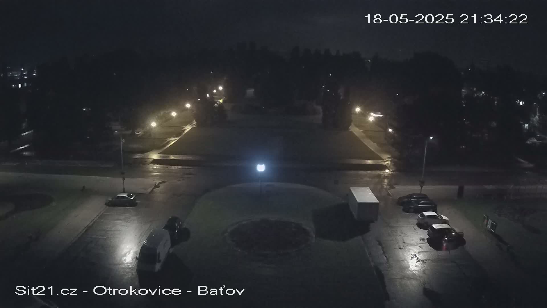 A nighttime, wet parking lot with several cars and a van, surrounded by dark trees and illuminated by streetlights.