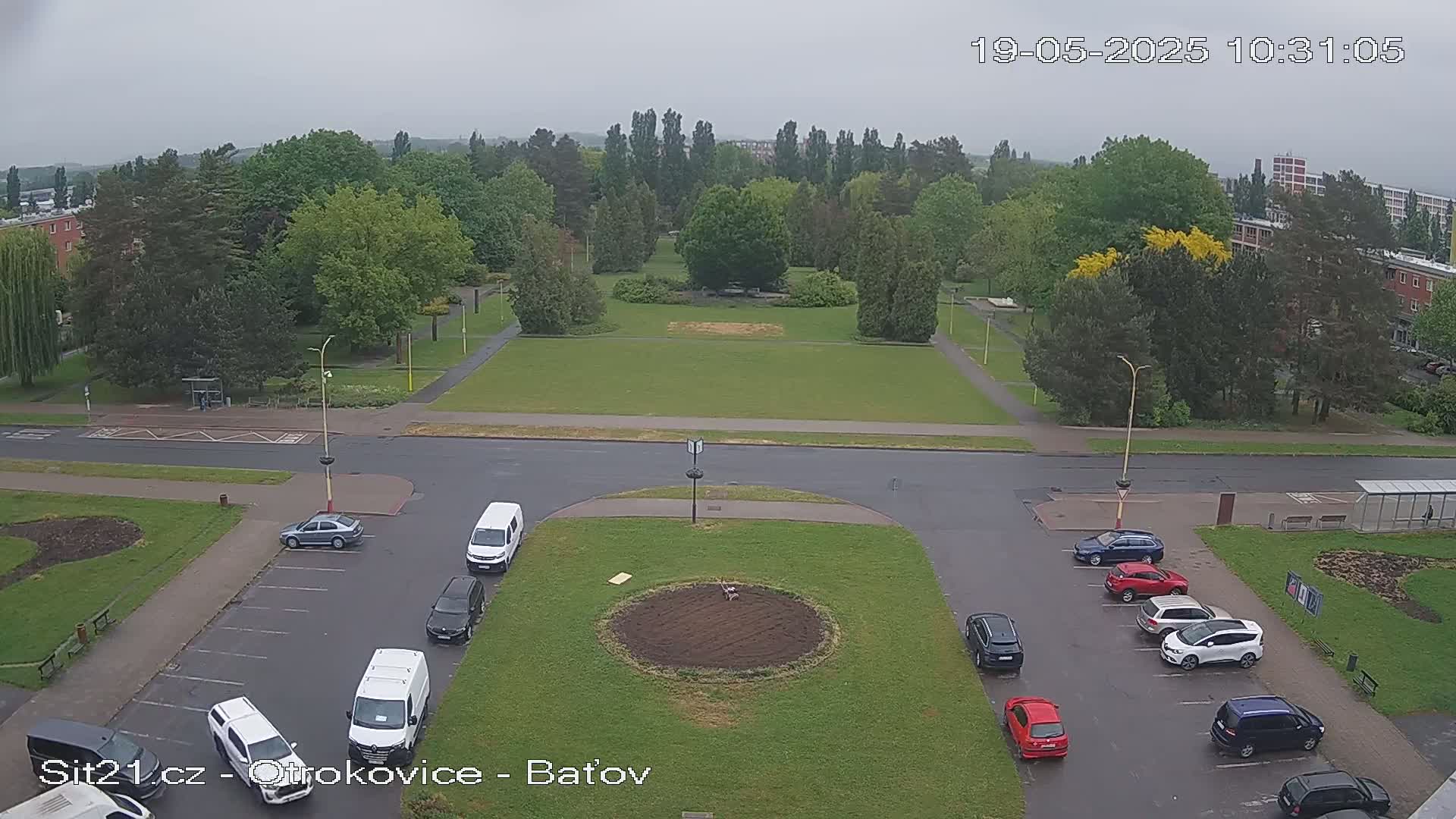 An overcast day reveals a high-angle view of a park and parking lot with several cars, surrounded by buildings and lush green trees.