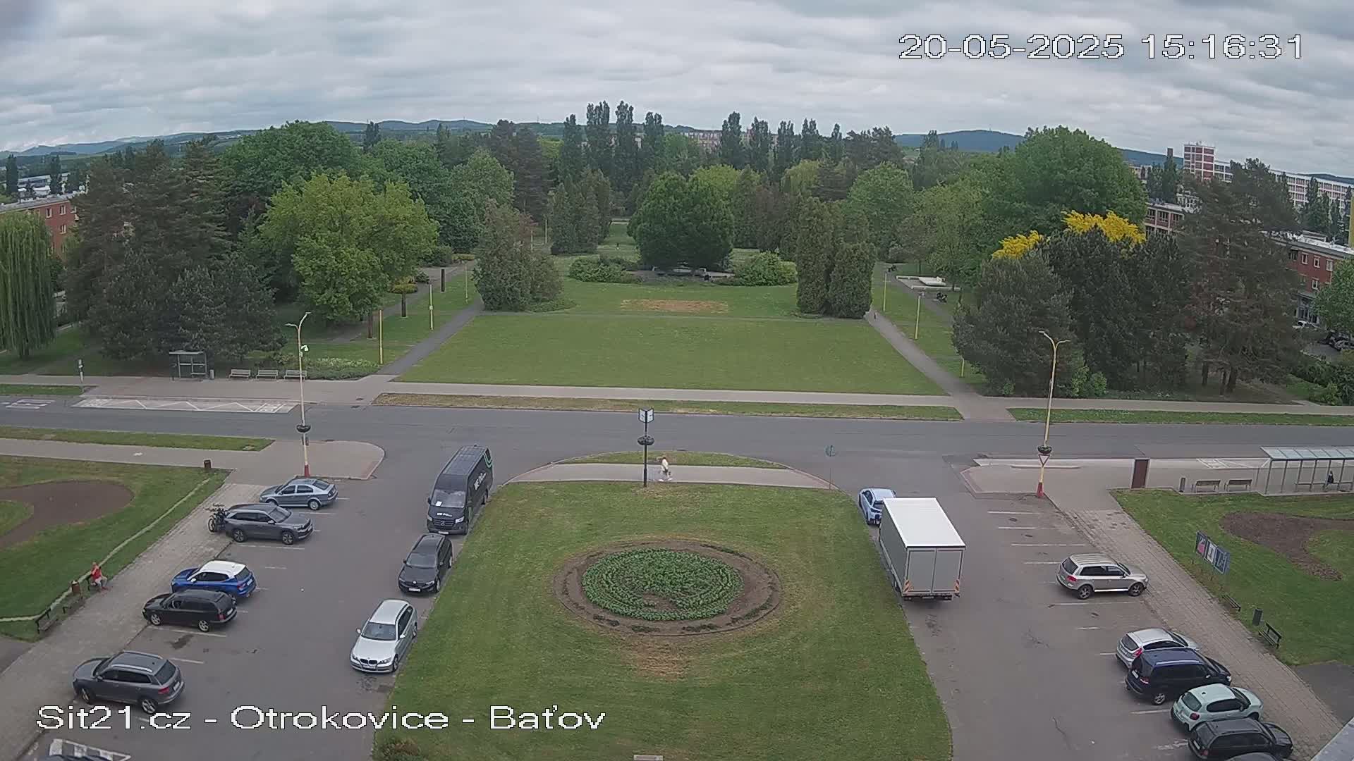 An overcast day reveals a park with a circular garden, surrounded by a street with parked cars and buildings in the background.