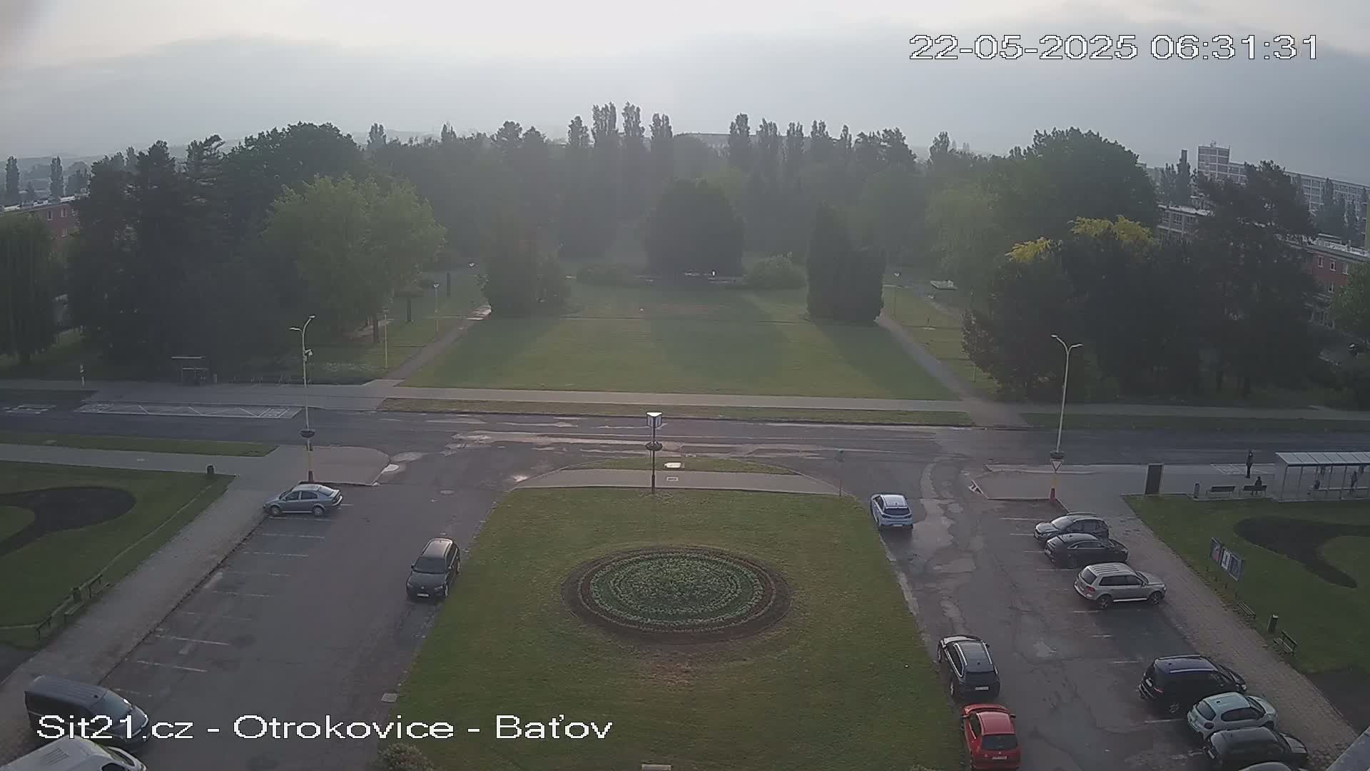 A misty morning scene shows a park with a circular flower bed in the center, surrounded by a roadway and parking lots with several cars.