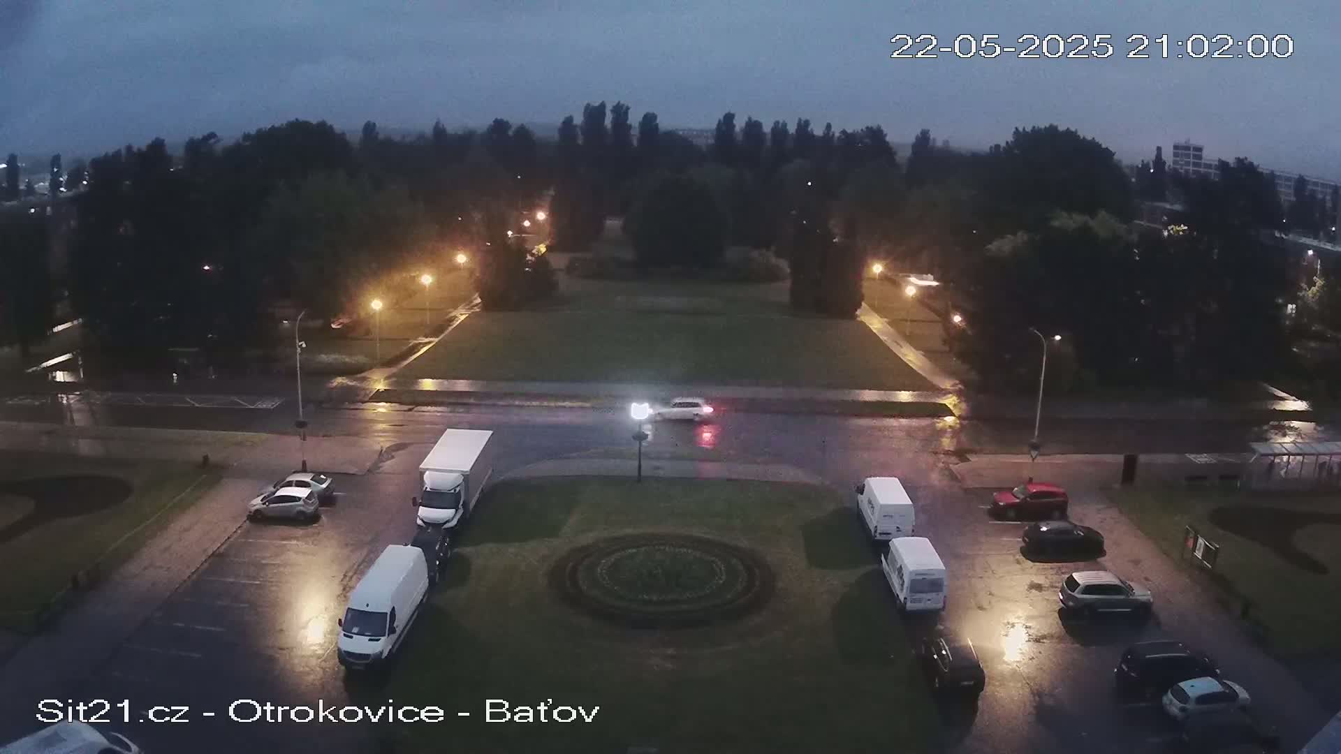 A wet, nighttime aerial view shows a park with a circular garden in the center, surrounded by a road with parked cars and vans, under a dark, overcast sky.