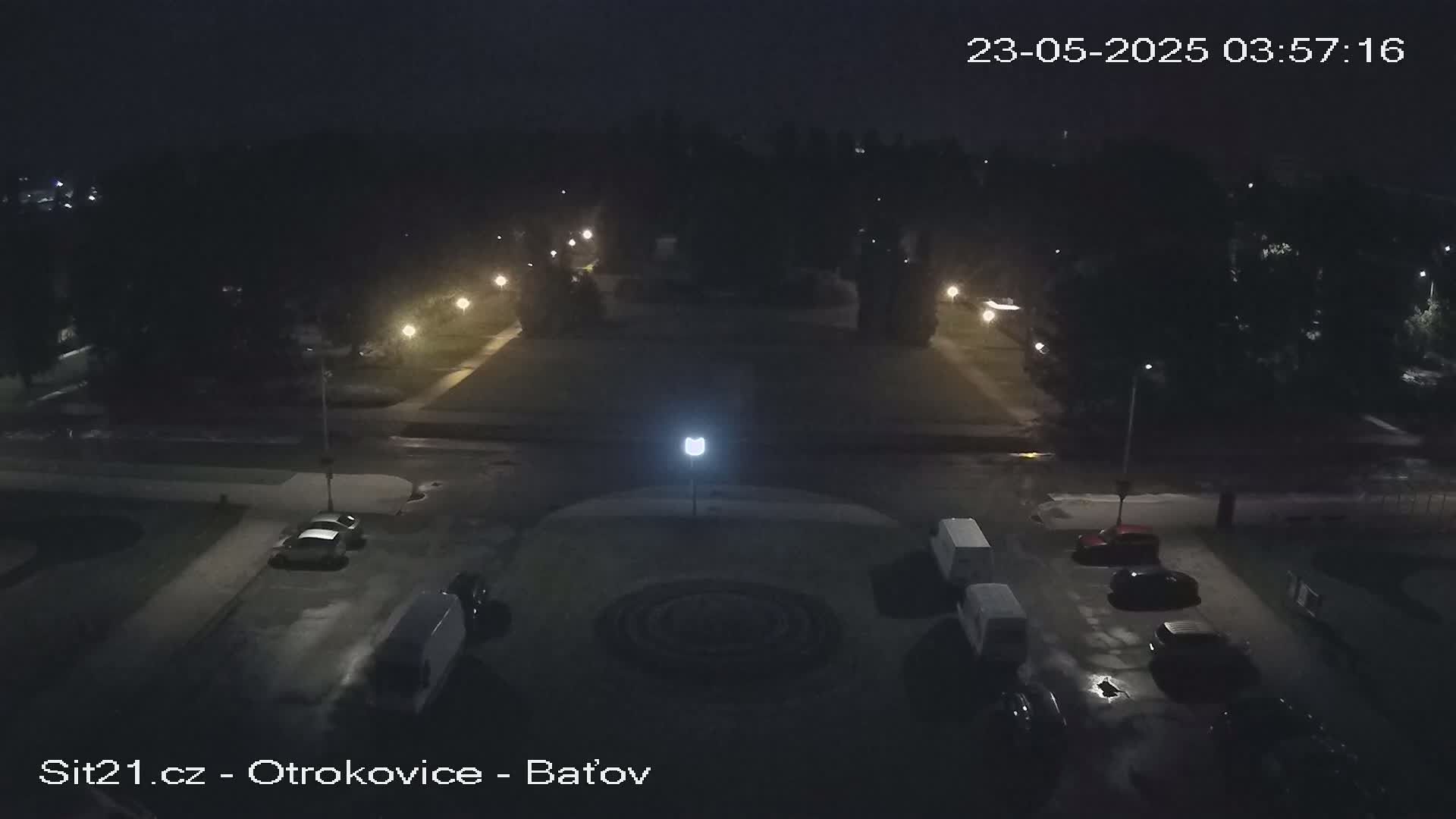 A nighttime, overhead view of a square plaza with several cars parked along the sides, surrounded by dark trees and illuminated by streetlights.