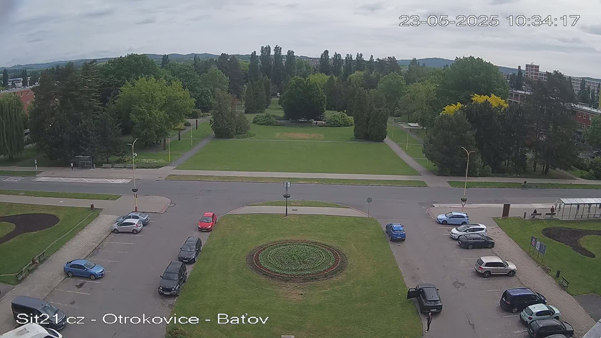 An overhead view of a park with a circular flower bed in the center, surrounded by a parking lot with several cars parked, under a partly cloudy sky.